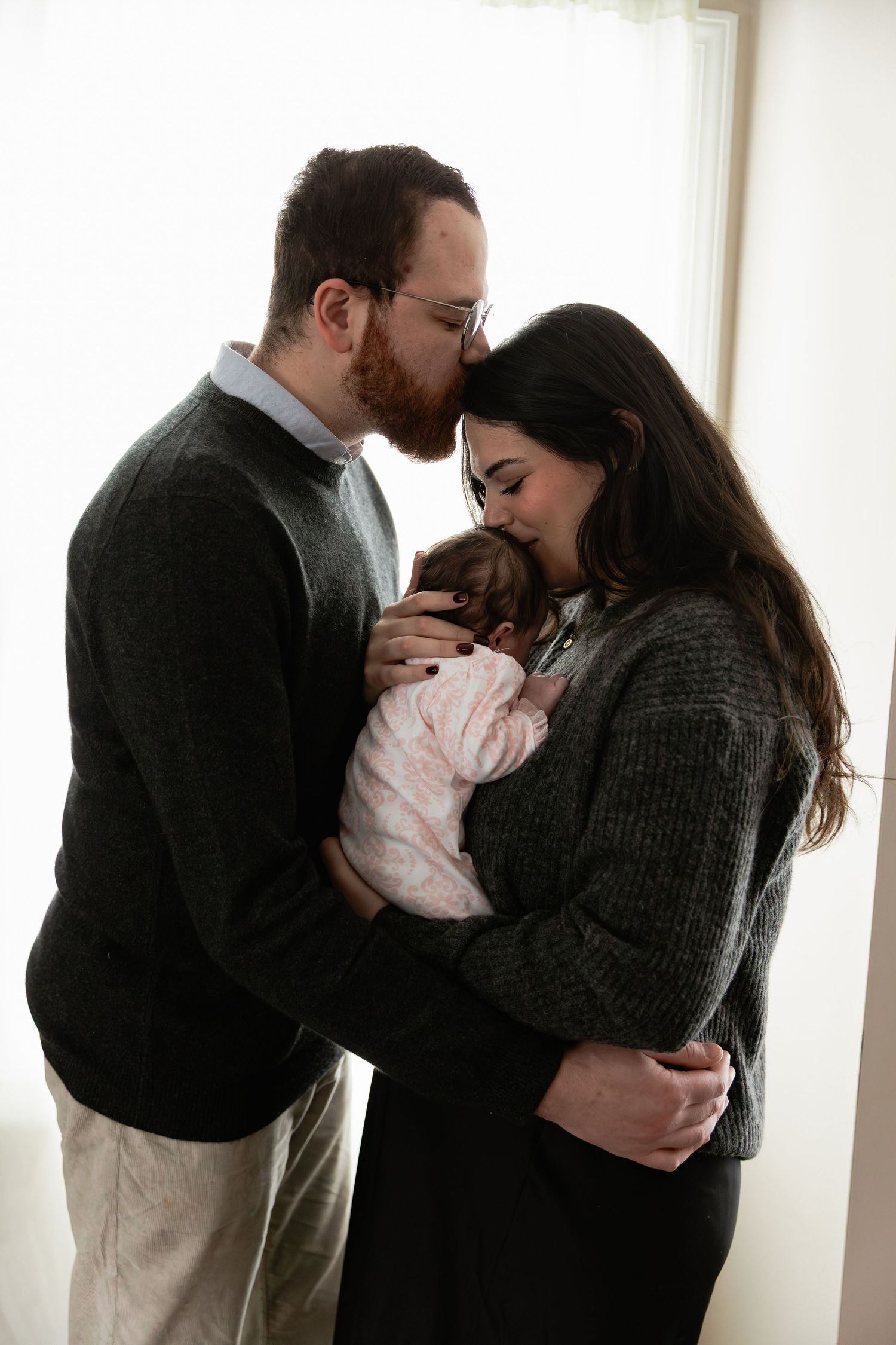 A couple in dark clothing embraces their newborn baby in three similar poses by a bright window.