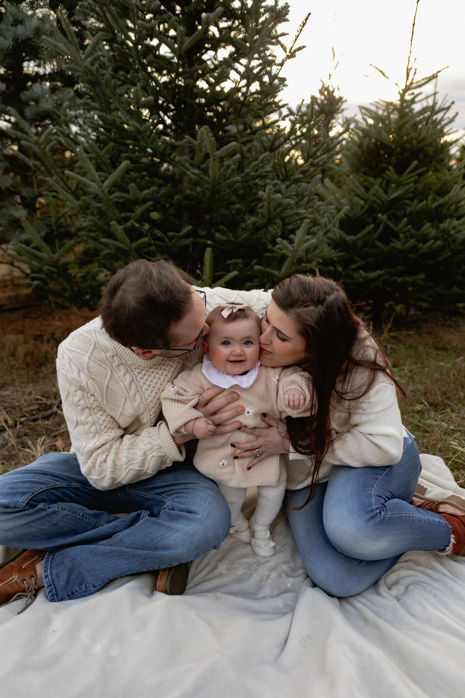 A family sits on a white blanket in front of evergreen trees, sharing kisses and embraces during a holiday photoshoot.