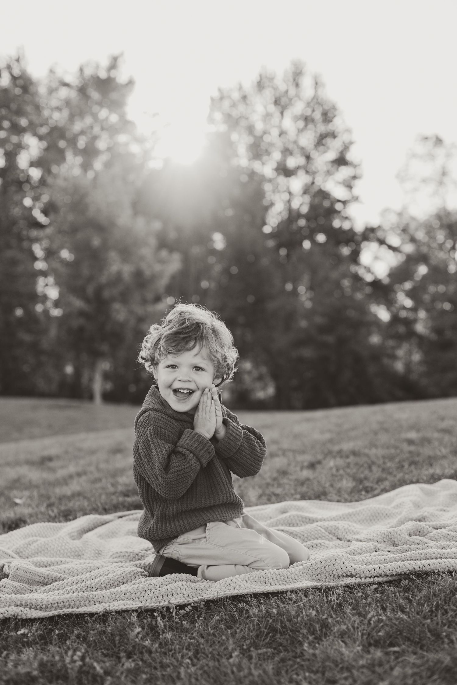 A child lies on a blanket outdoors in black and white, laughing and covering their face during a sunset photoshoot.