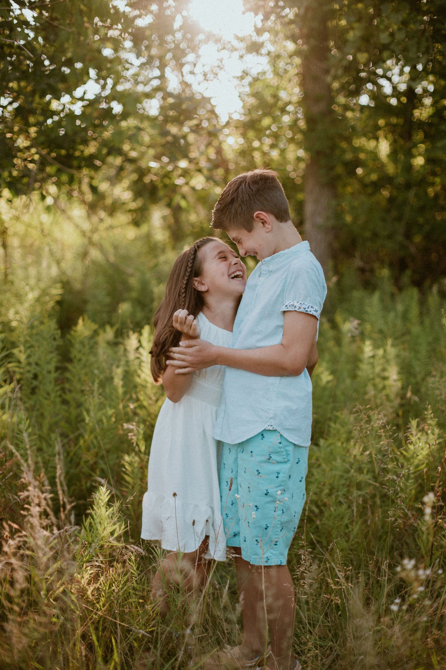 A romantic couple embraces playfully in a sunlit meadow with tall grass and golden hour lighting.