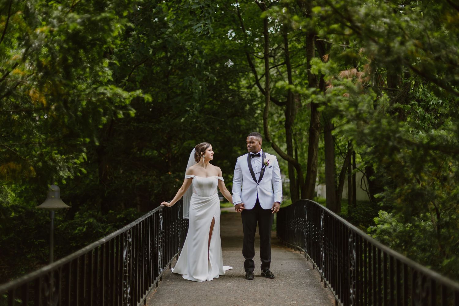 A couple in wedding attire walks hand in hand down a wooden bridge path surrounded by lush green forest.