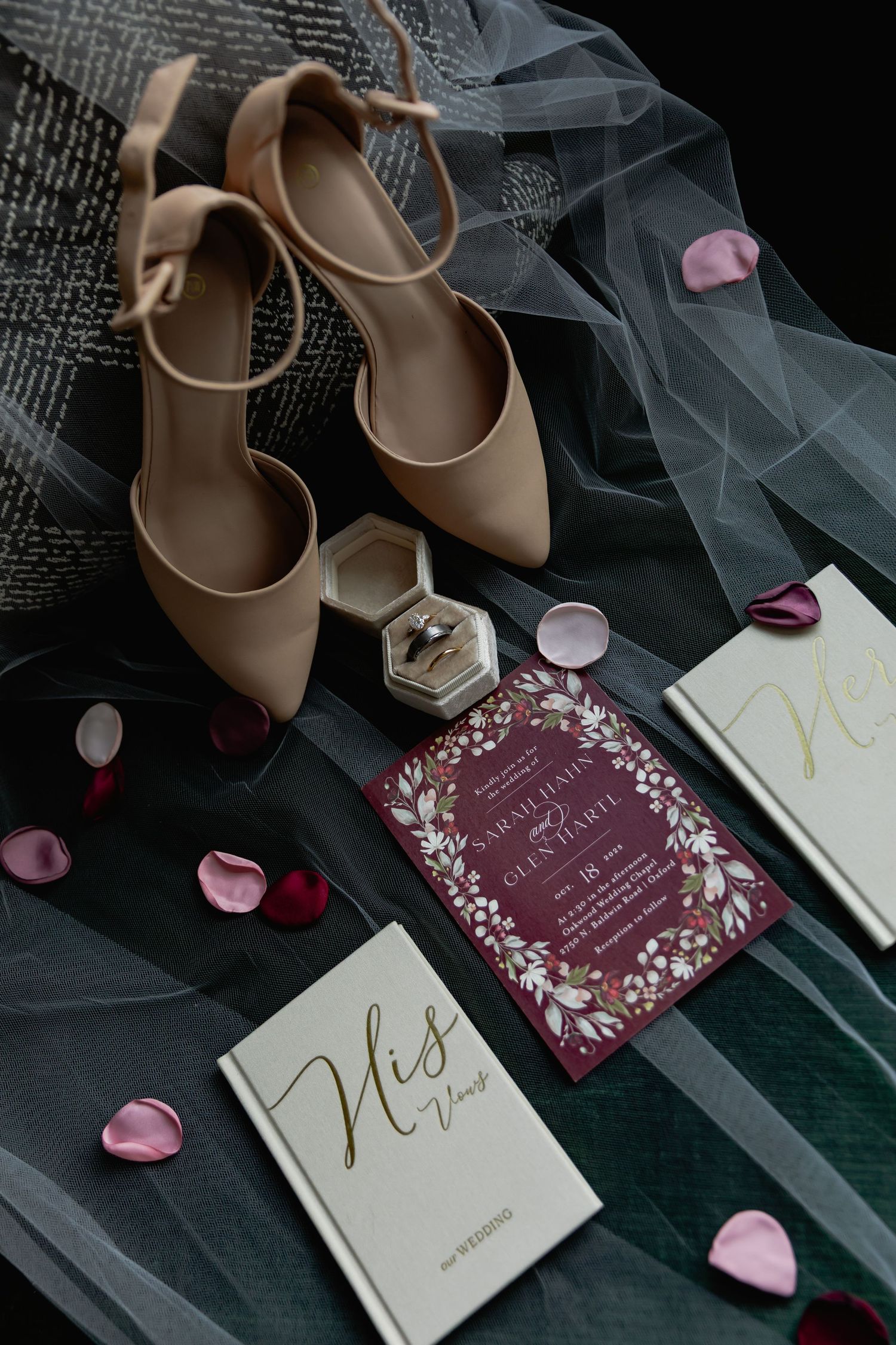 Wedding details flatlay with nude heels, veil, invitation cards, ring box and scattered pink rose petals on dark background.