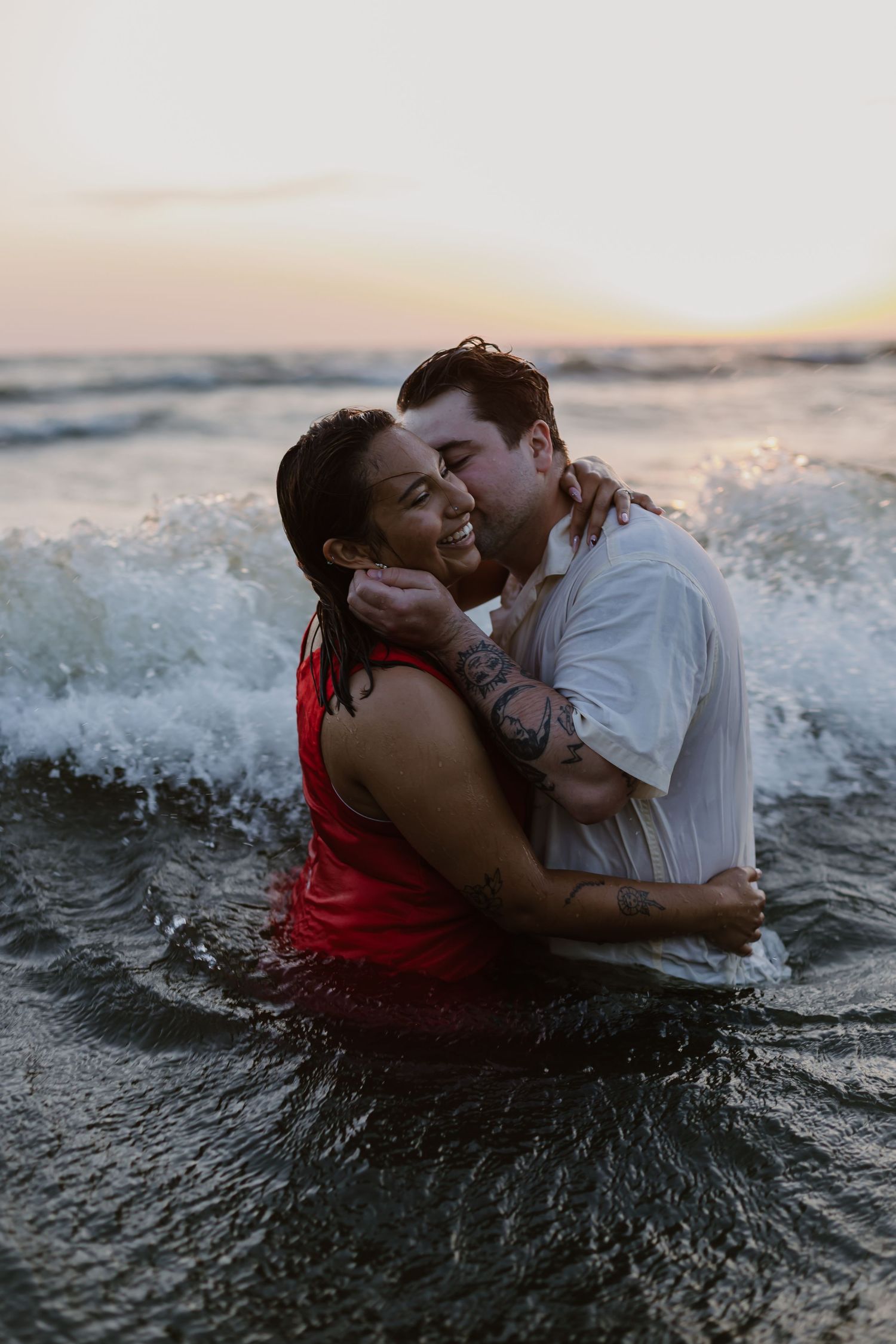 A couple embraces and kisses while sitting in ocean waves during sunset, creating a romantic silhouette against the water.