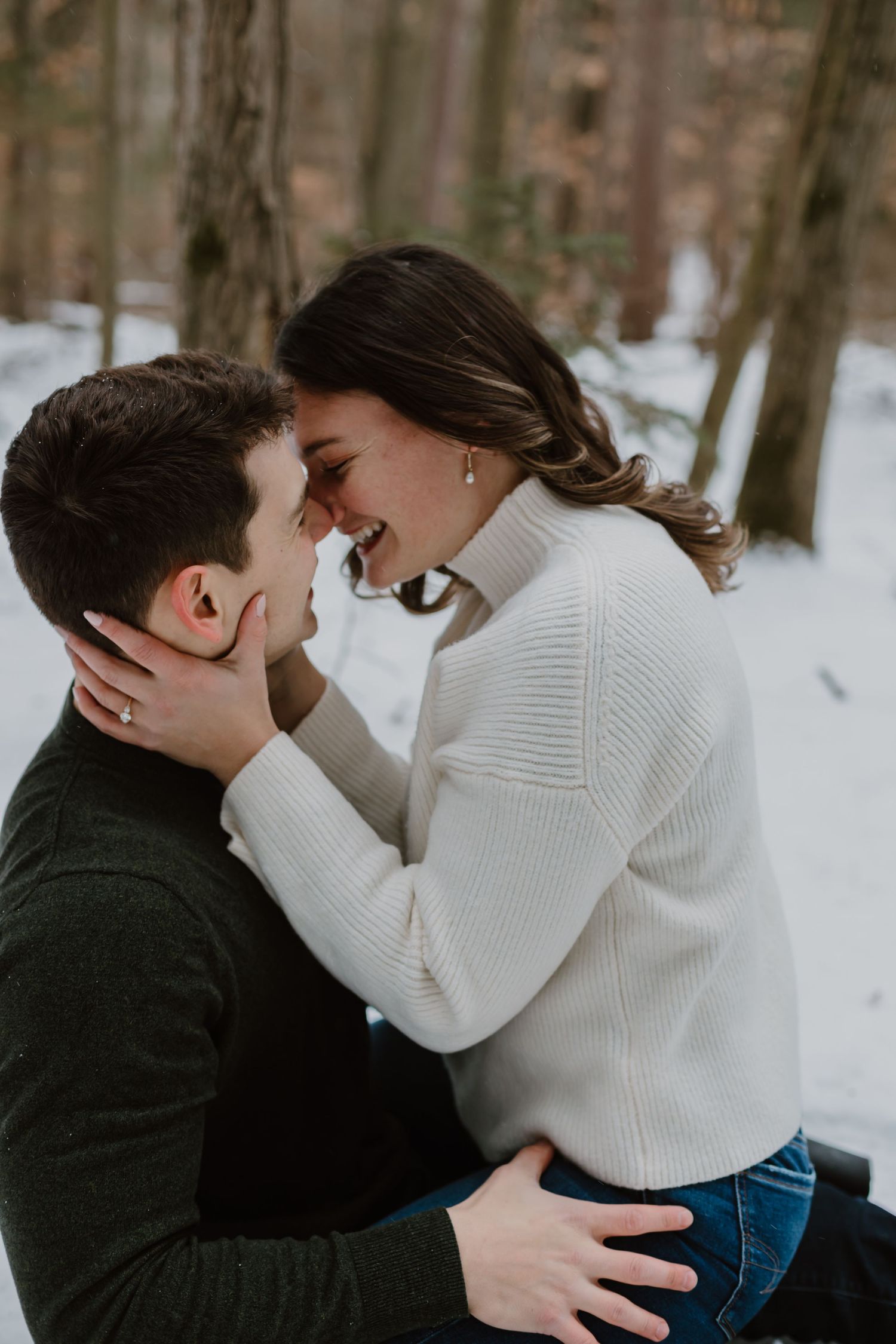 Romantic winter engagement photos show couple in white sweater and dark jacket embracing in snowy forest setting.