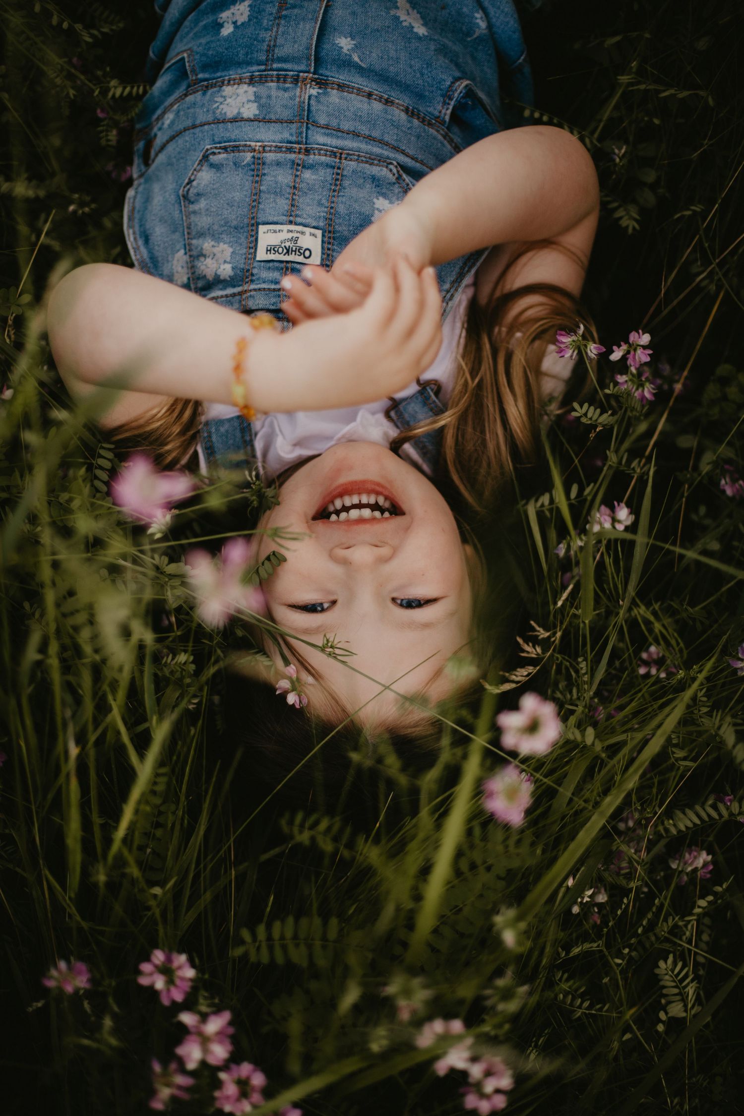 Someone lying in green grass surrounded by delicate pink wildflowers wearing denim overalls on a sunny day.