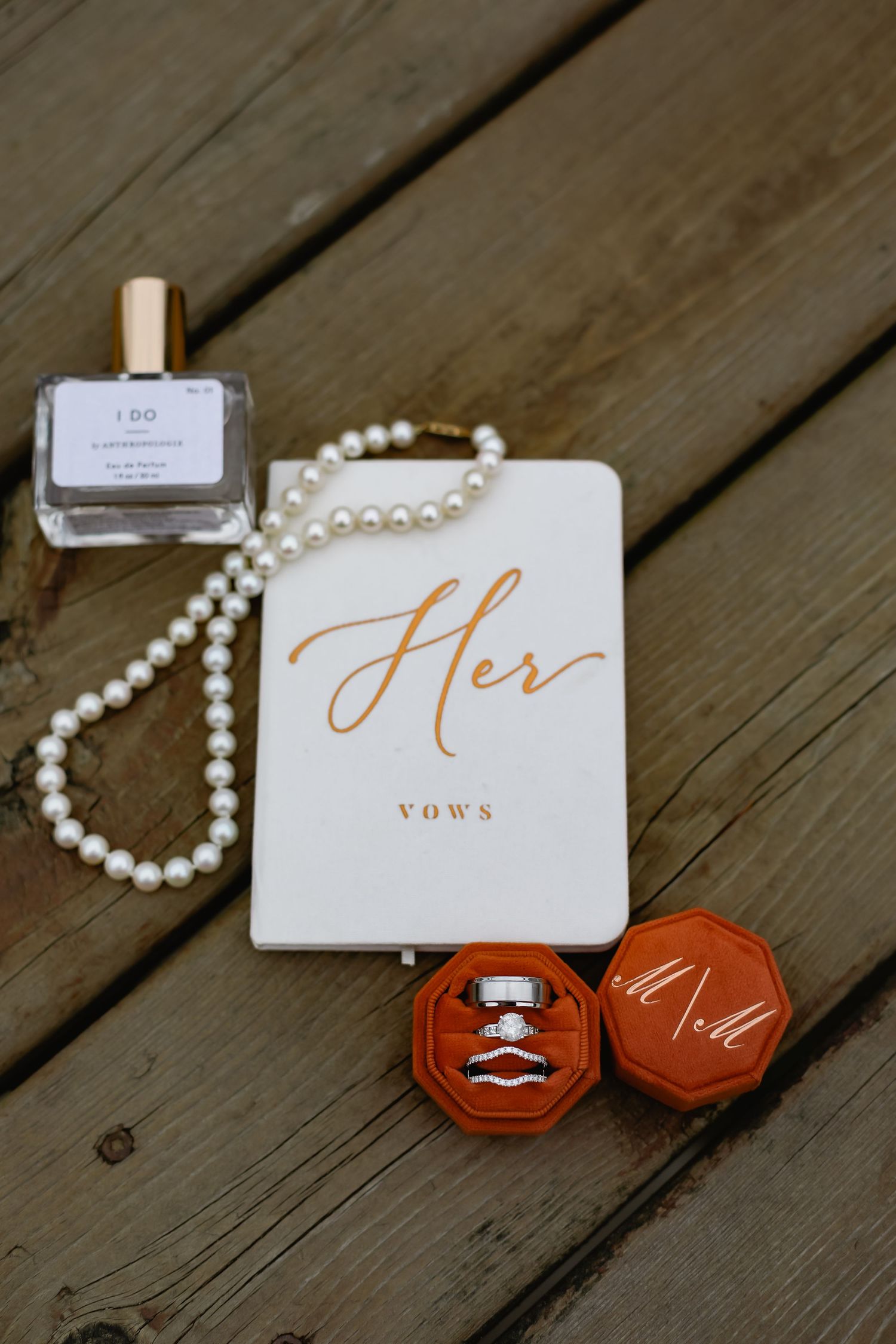 Wedding vows book with pearl necklace and red ring boxes arranged on rustic wooden deck.