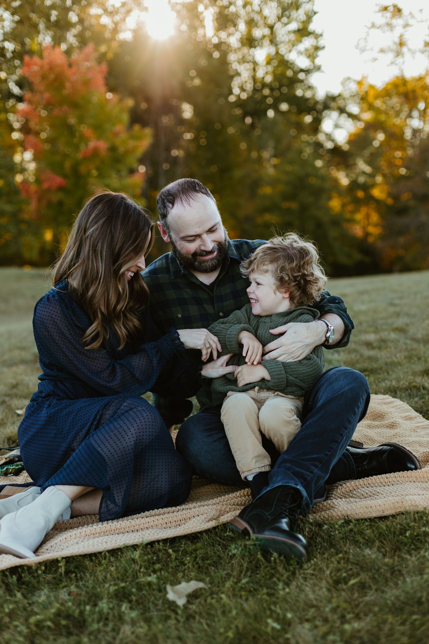 A happy family laughs while snuggling together on a blanket during a golden autumn sunset in a grassy field.