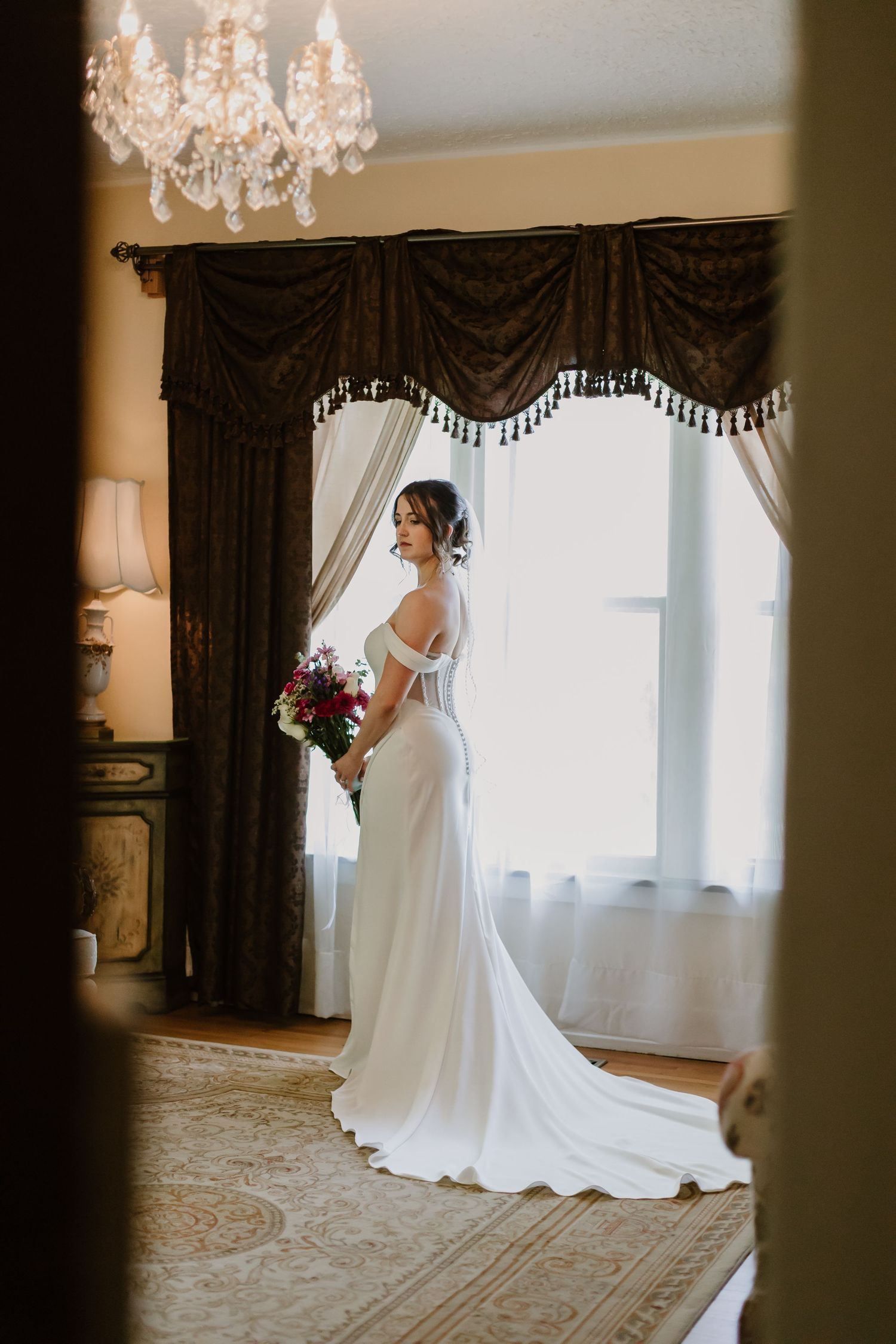 A bride in a flowing white wedding dress poses gracefully in an elegant room with dark curtains and chandeliers.
