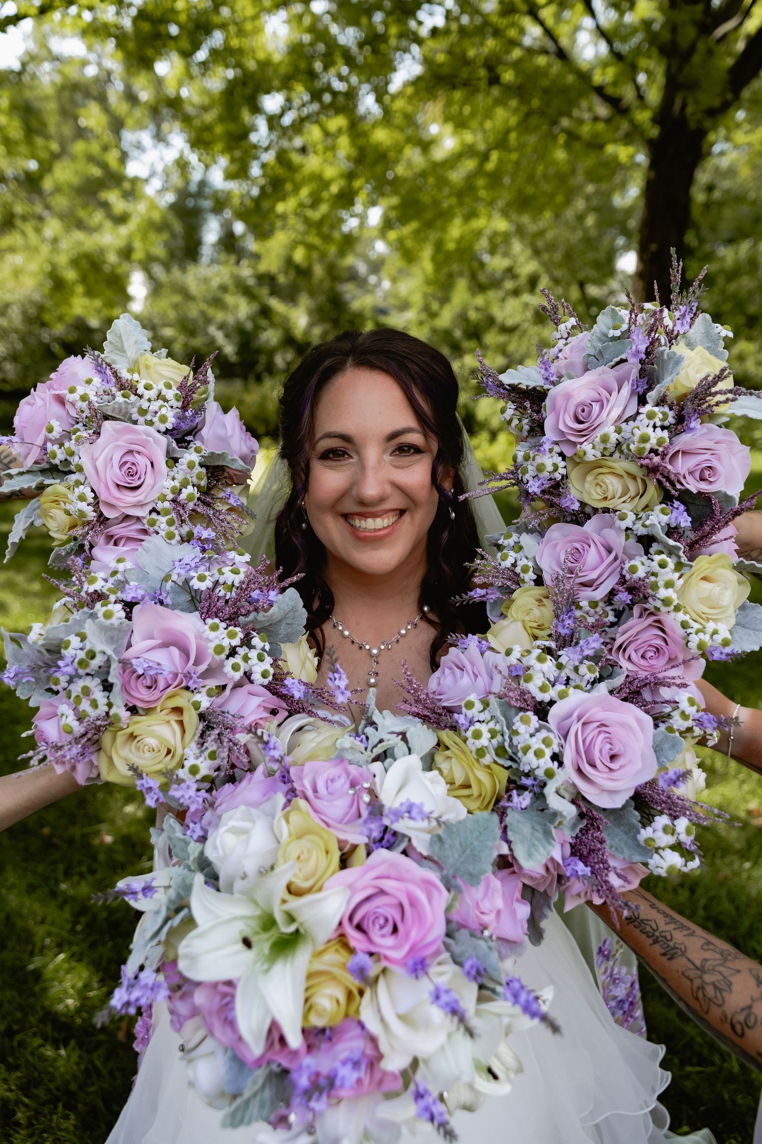 Stunning purple and white floral wedding bouquet with roses and lilies against green outdoor background.