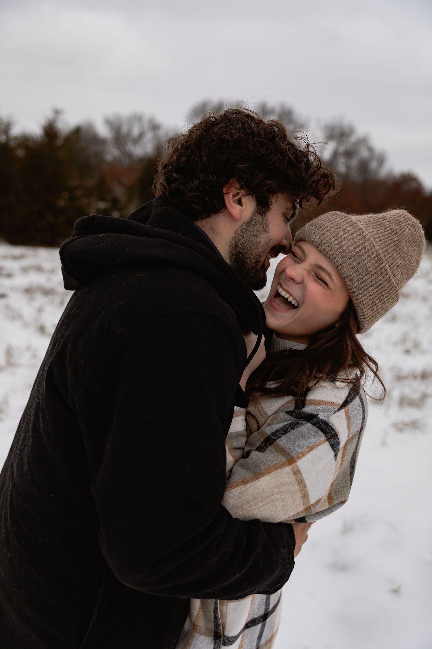A couple shares joyful laughter while embracing in a snowy winter landscape with bare trees in the background.