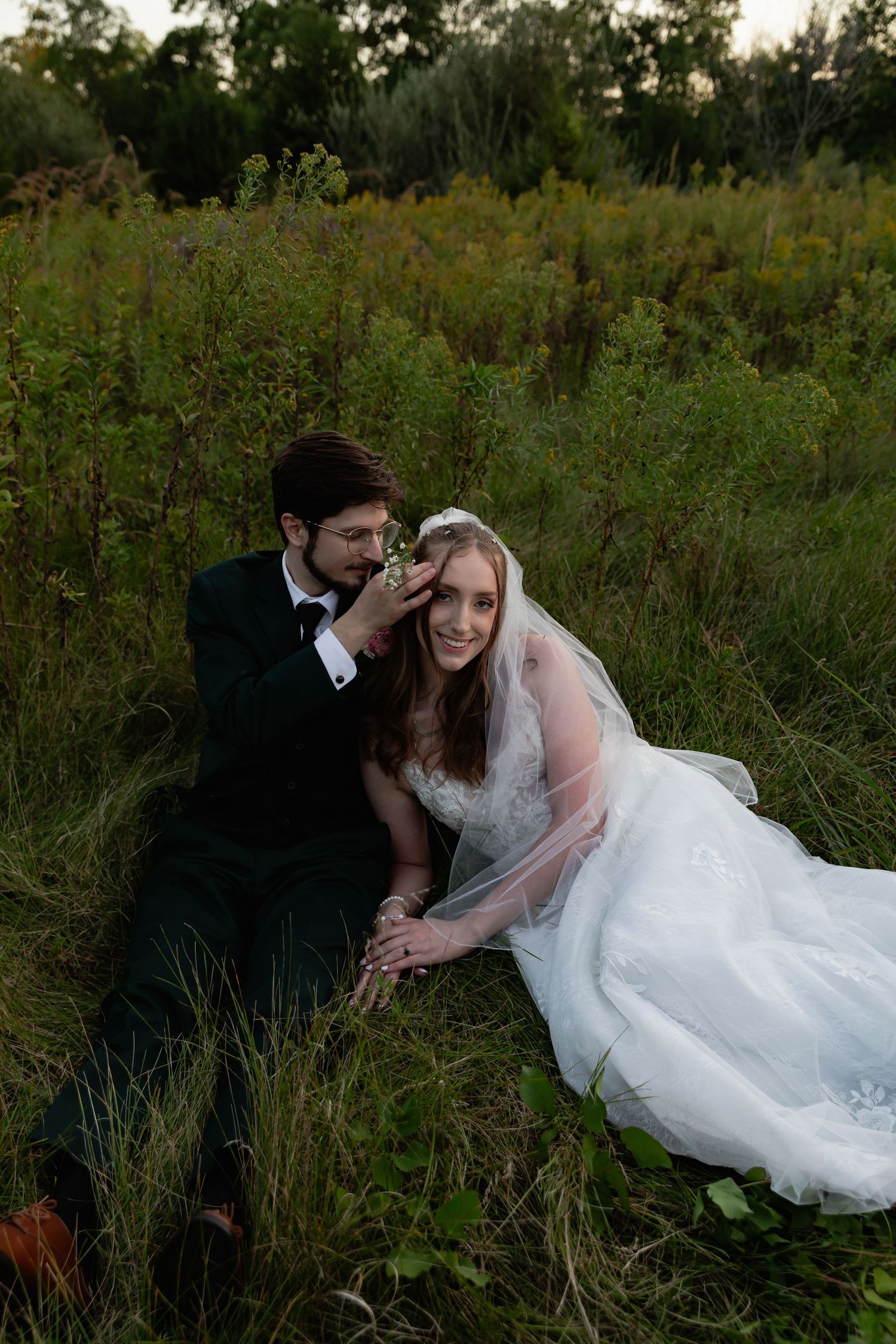 A romantic wedding photo of a couple sitting together in a lush green meadow at sunset.