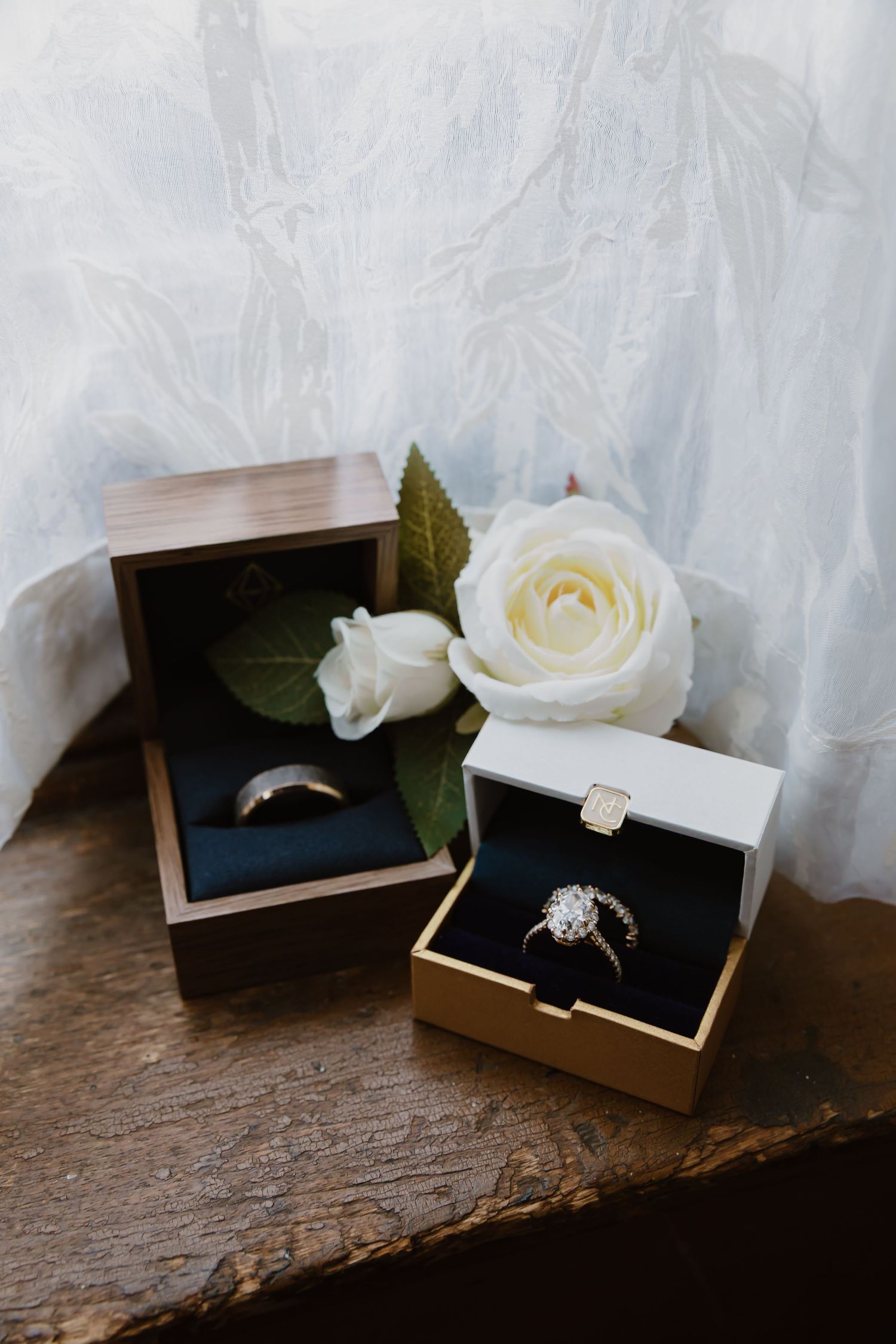 Elegant diamond engagement ring displayed in dark wooden box with white roses on rustic wooden surface.