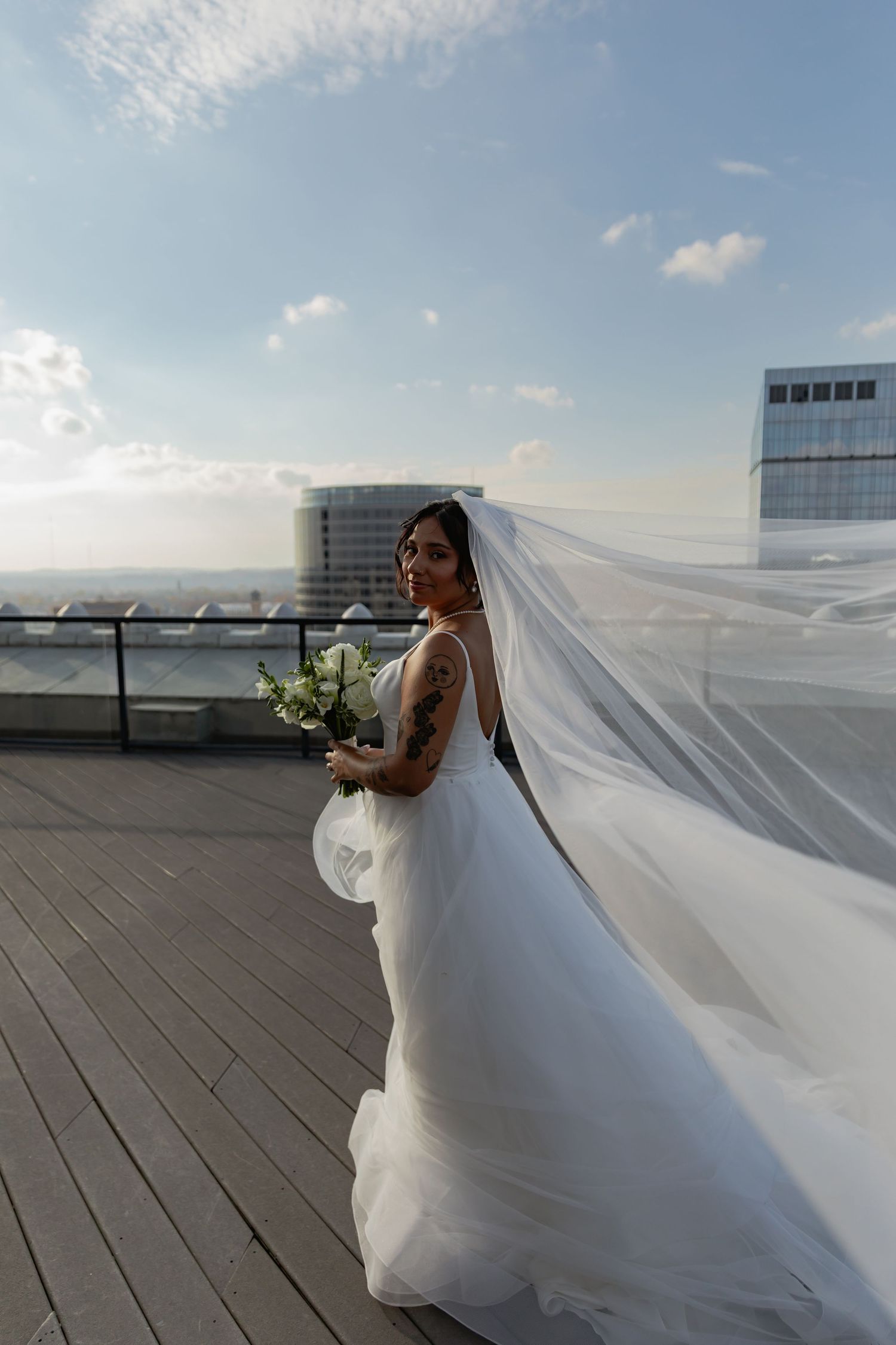 A flowing white wedding veil catches the wind dramatically against a rooftop cityscape backdrop.