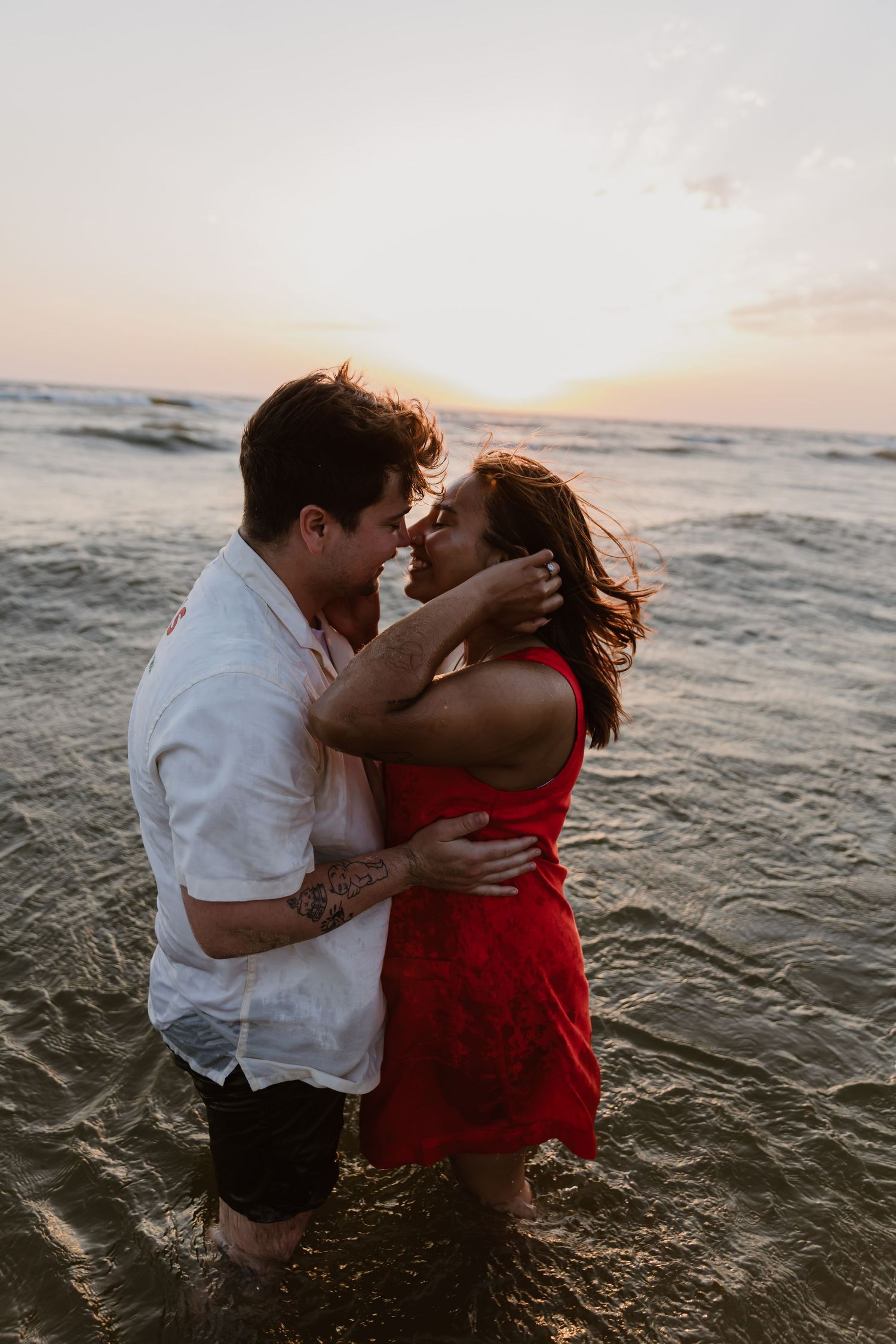 A couple embraces while standing in ocean waves at sunset, with one wearing a red dress and the other in white shirt.