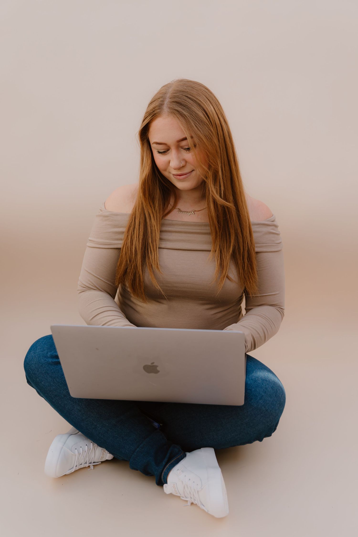 Person in beige top and jeans working on MacBook laptop while sitting cross-legged.
