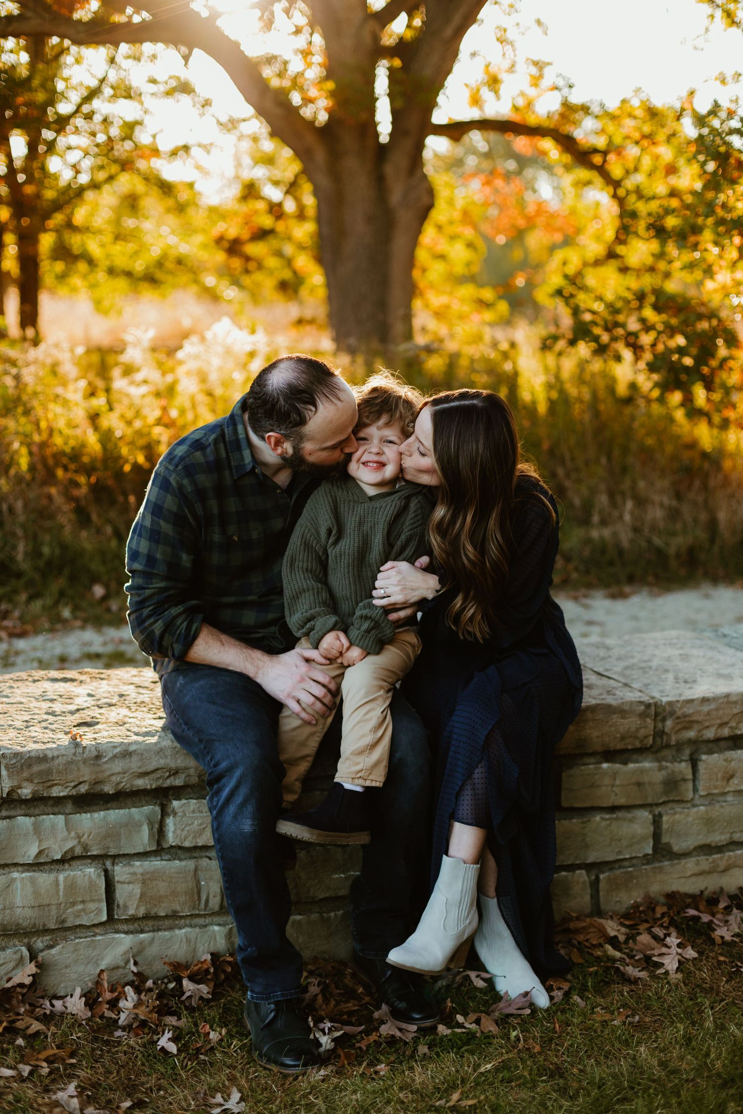 A couple sits together on a stone wall surrounded by autumn foliage during a romantic outdoor photoshoot at sunset.