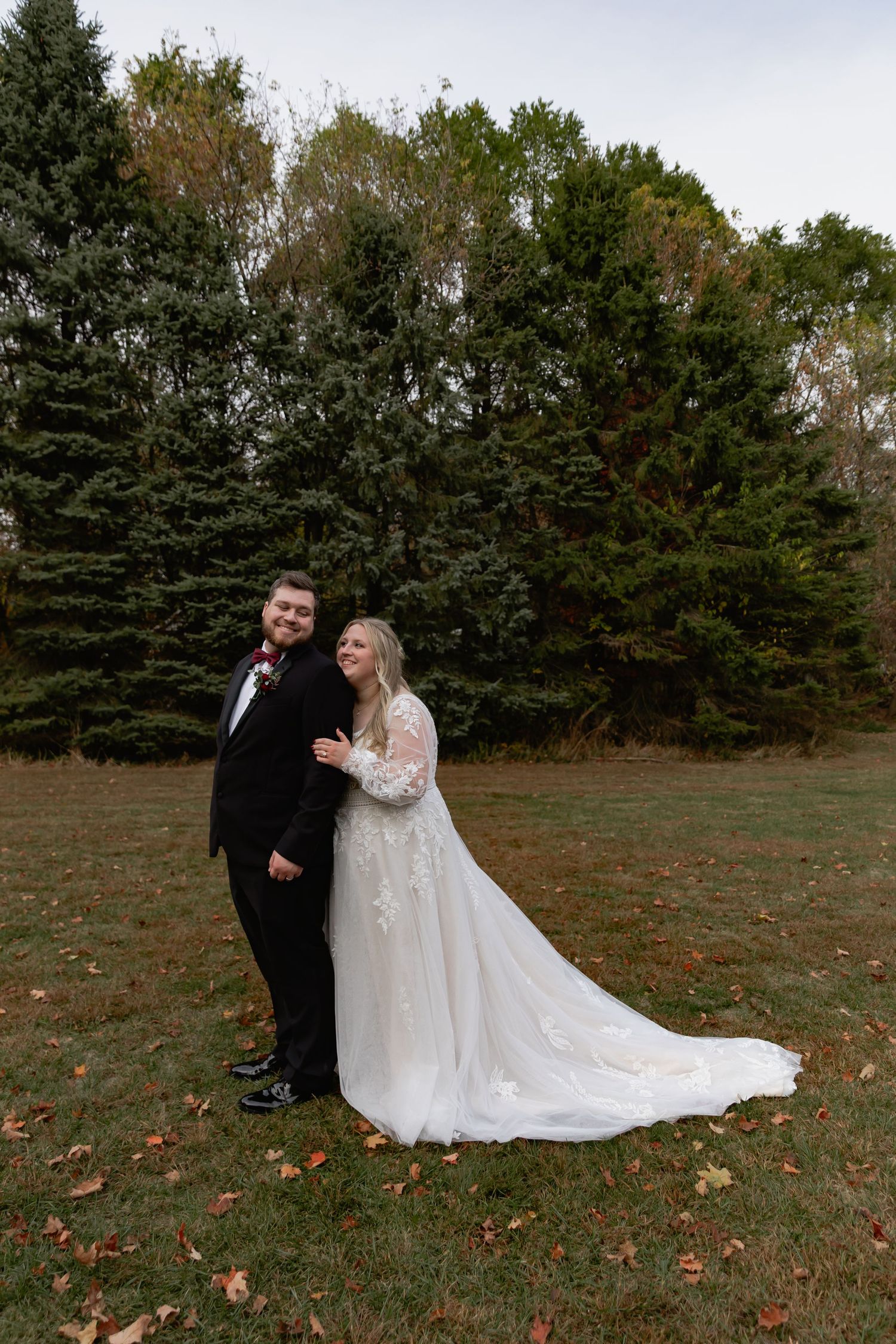 A couple in wedding attire poses together on a lawn with evergreen trees in the background during autumn.