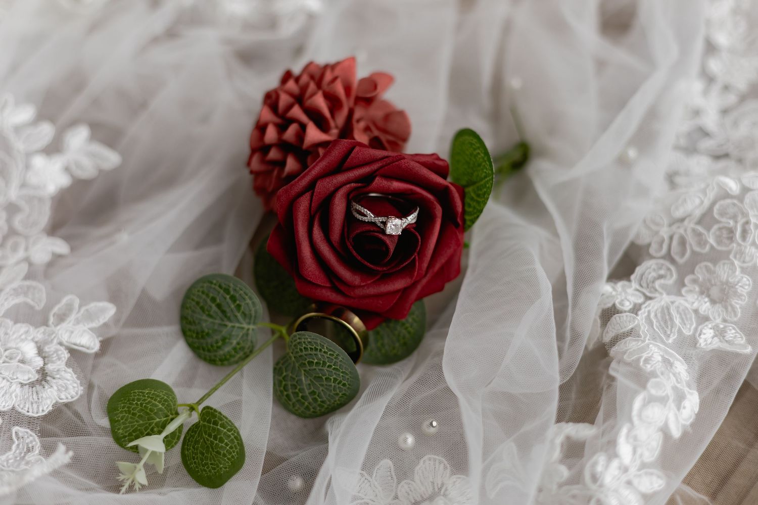 Wedding ring nestled inside a red rose bloom on white lace fabric with green leaves.