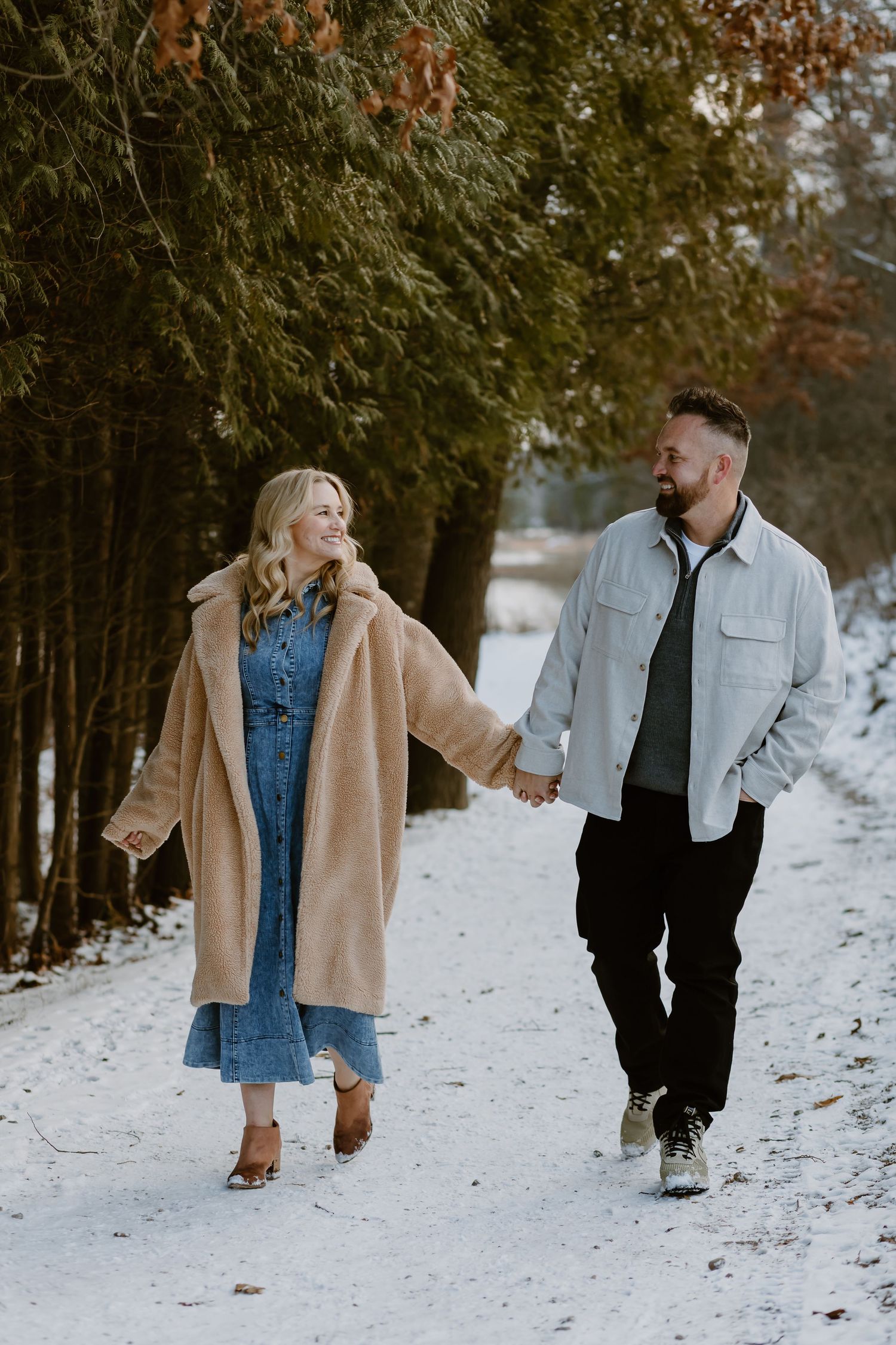 Couple walking hand in hand on snowy path with evergreen trees in background.