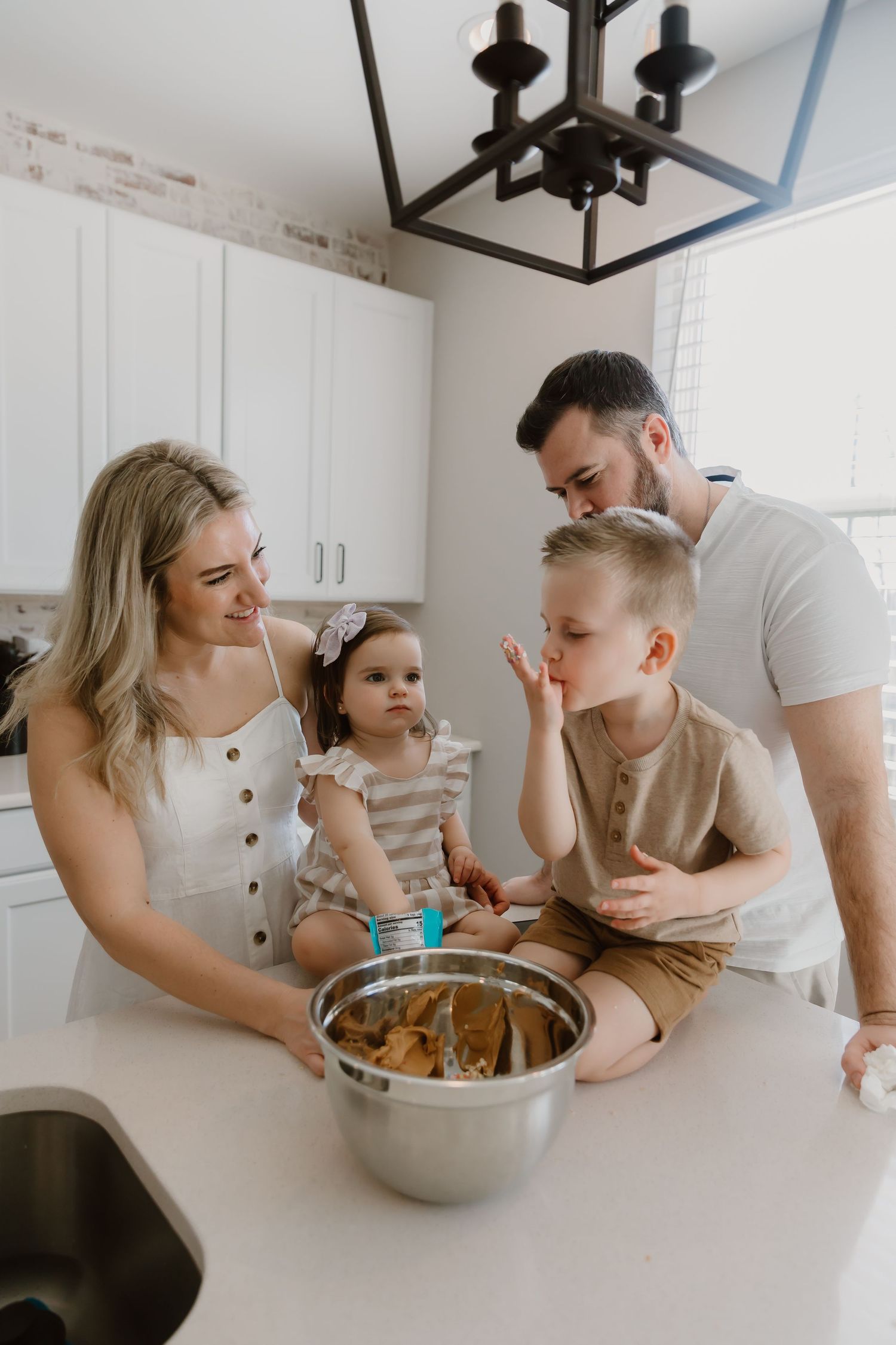 A family gathers around their kitchen counter while baking together in a modern white kitchen with pendant lighting.