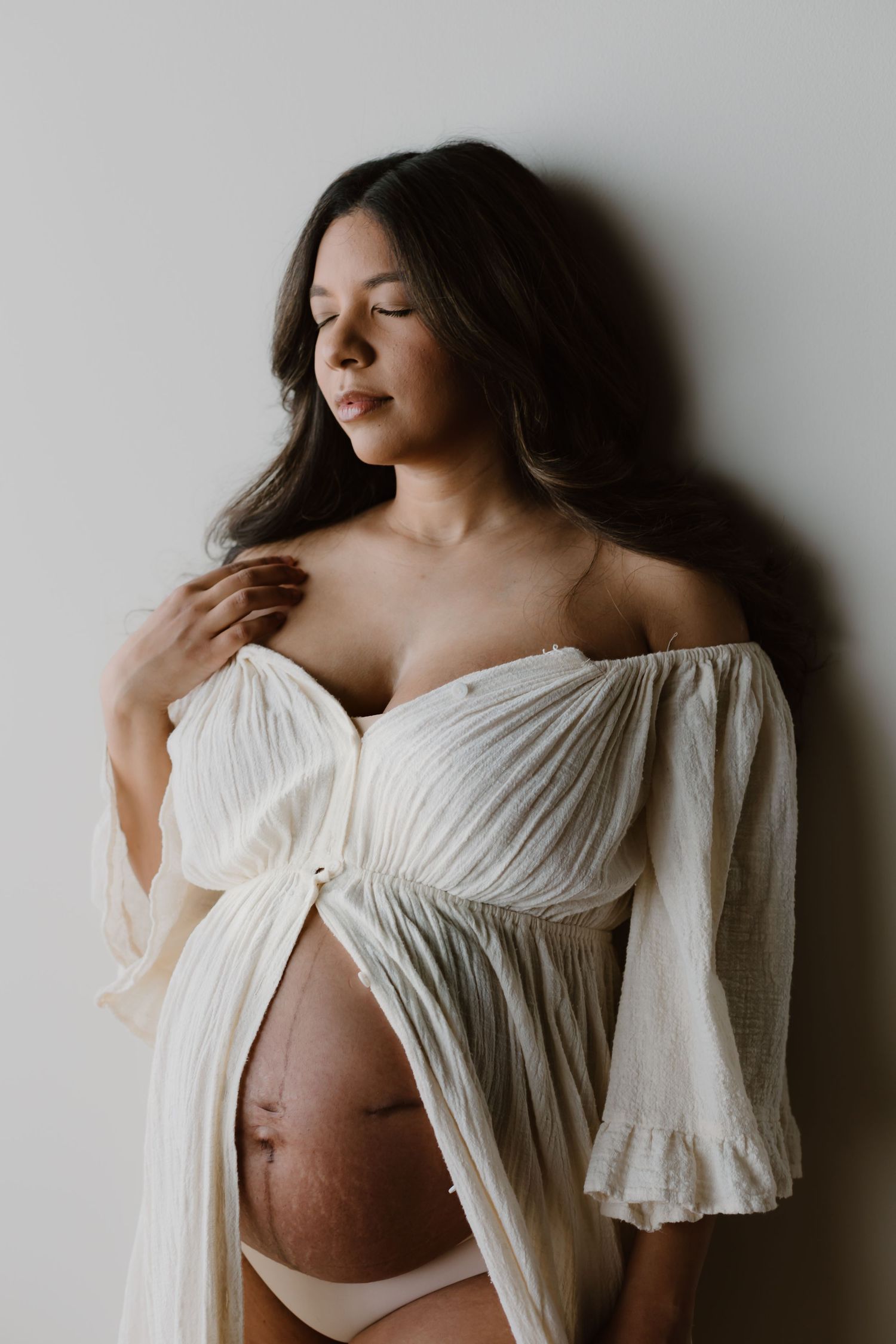 Maternity portrait showing a serene moment in a flowing white off-shoulder dress against neutral background.