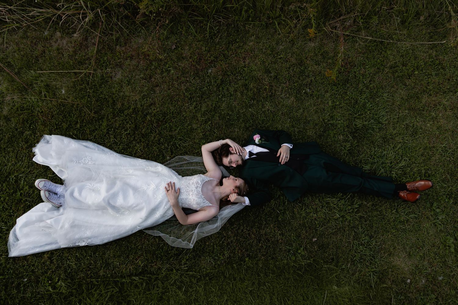 A bride and groom lay on green grass during a wedding photo session, captured from above.