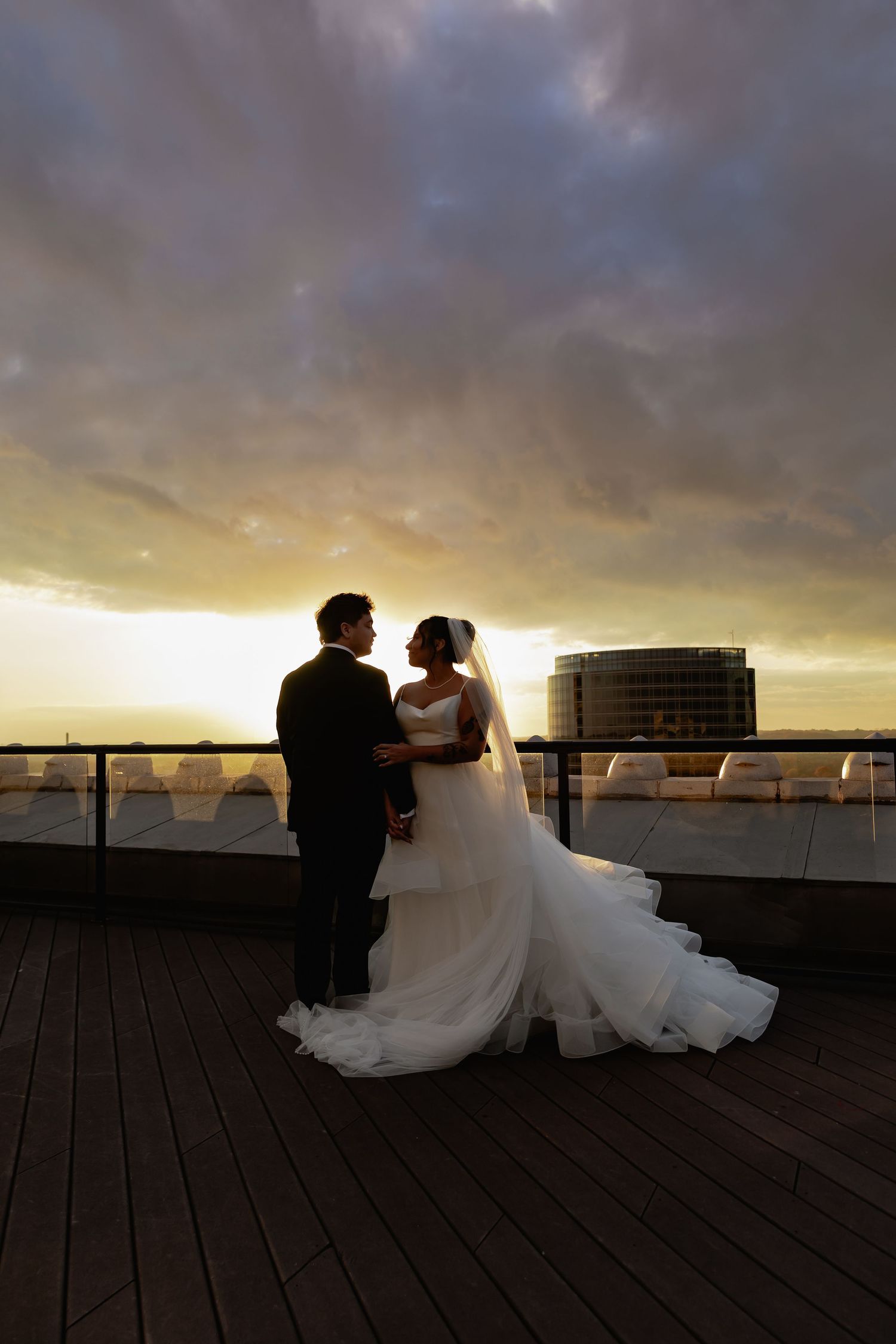 A romantic silhouette of a couple in formal attire embracing on a wooden deck at sunset with dramatic clouds overhead.