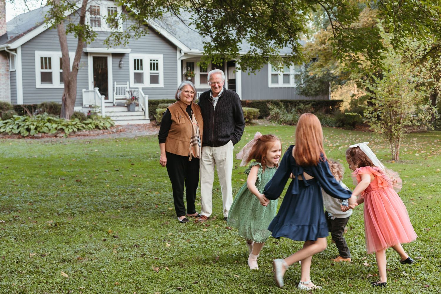Children in dresses dance together on a lawn while adults watch in the background.