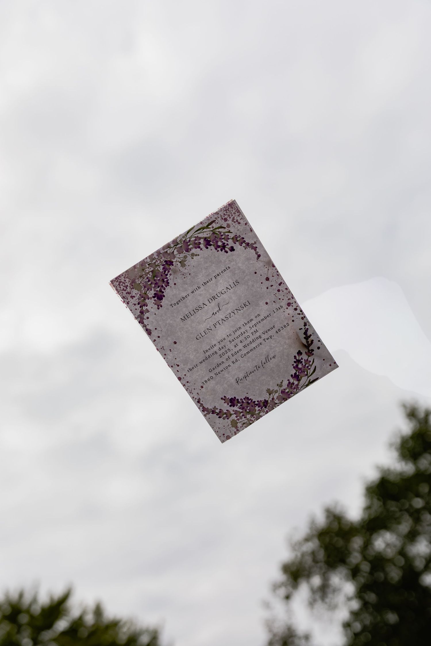 A wedding invitation with purple floral design appears to float against a cloudy sky.