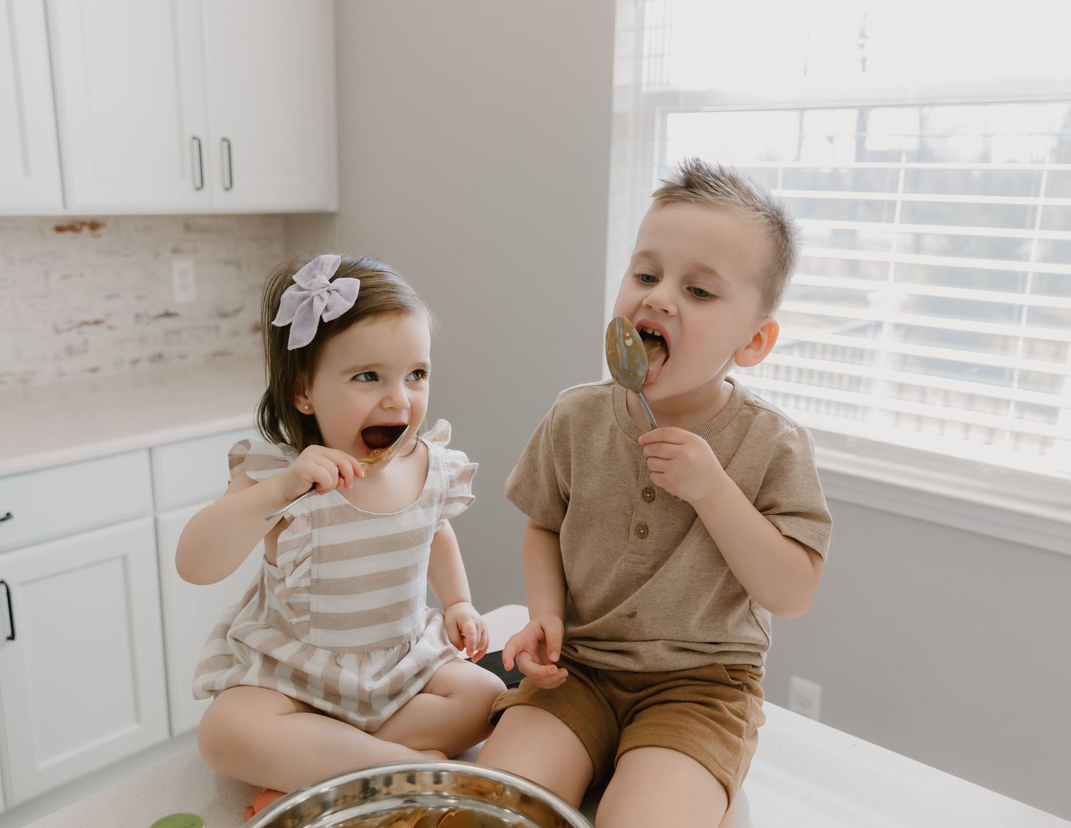 A child in a tan shirt eats a chocolate cookie while sitting in a bright white kitchen.