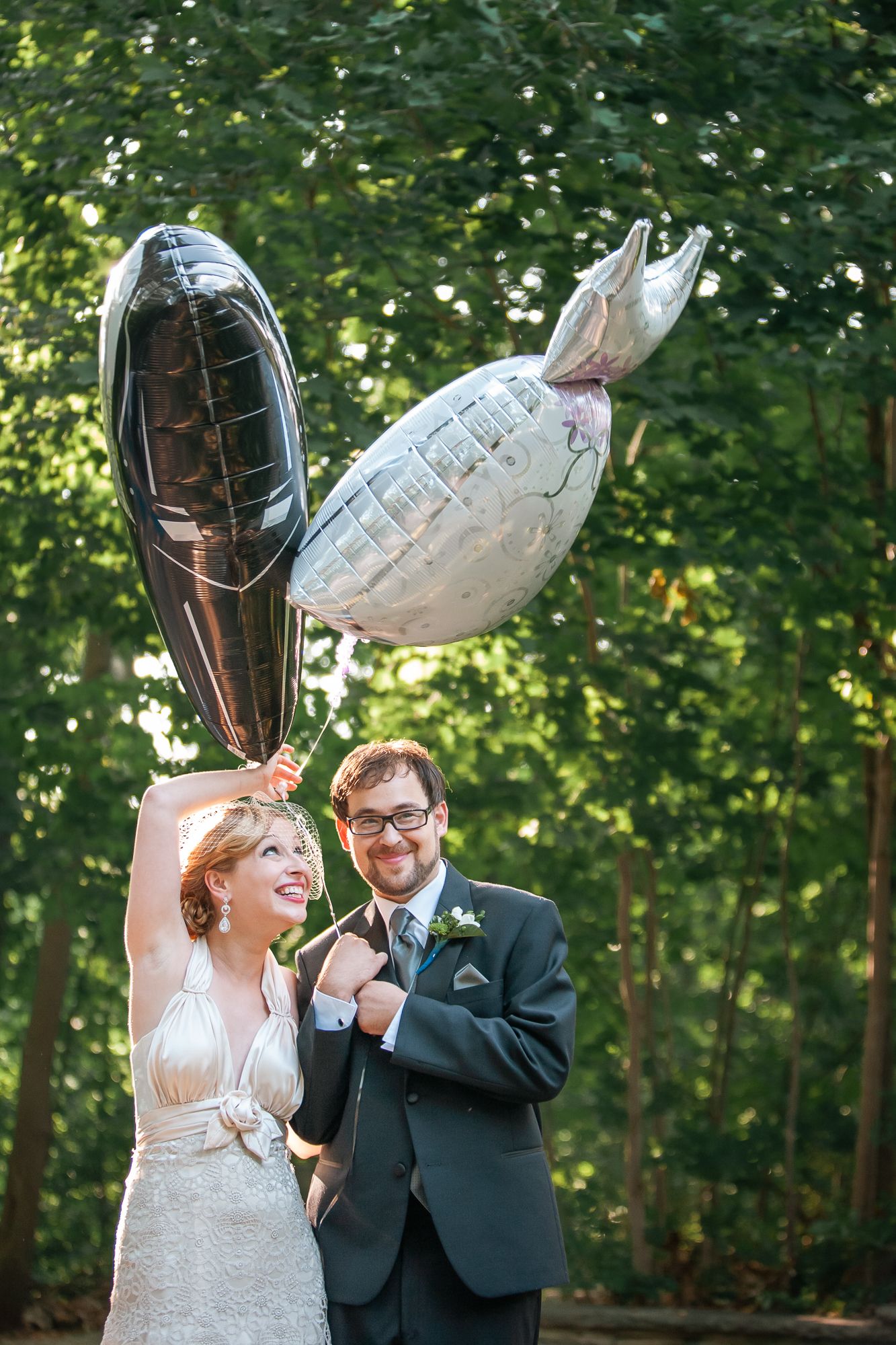 Happy couple in formal attire holding silver balloons outdoors in a garden setting during a celebration.
