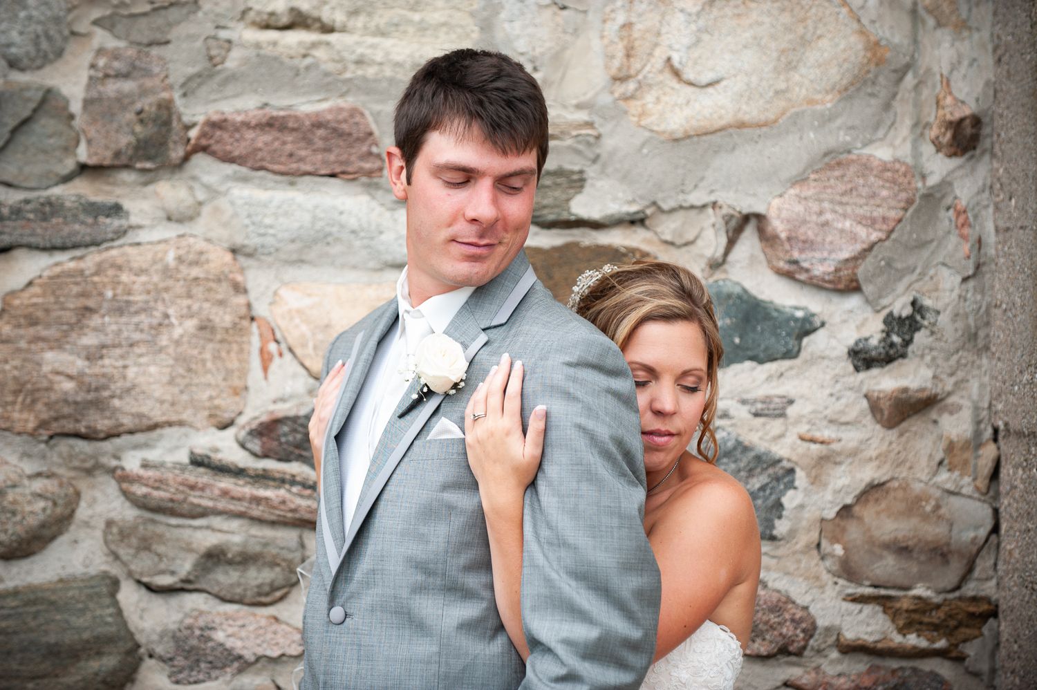 A bride and groom pose together against a rustic stone wall in formal wedding attire.