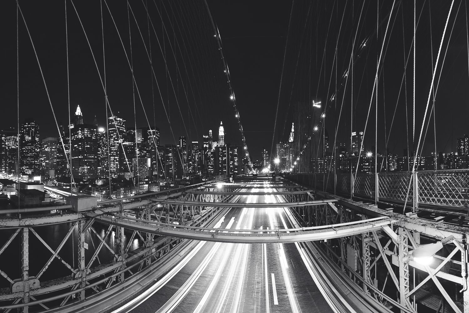 Long exposure night shot of light trails on Brooklyn Bridge with Manhattan skyline in background.