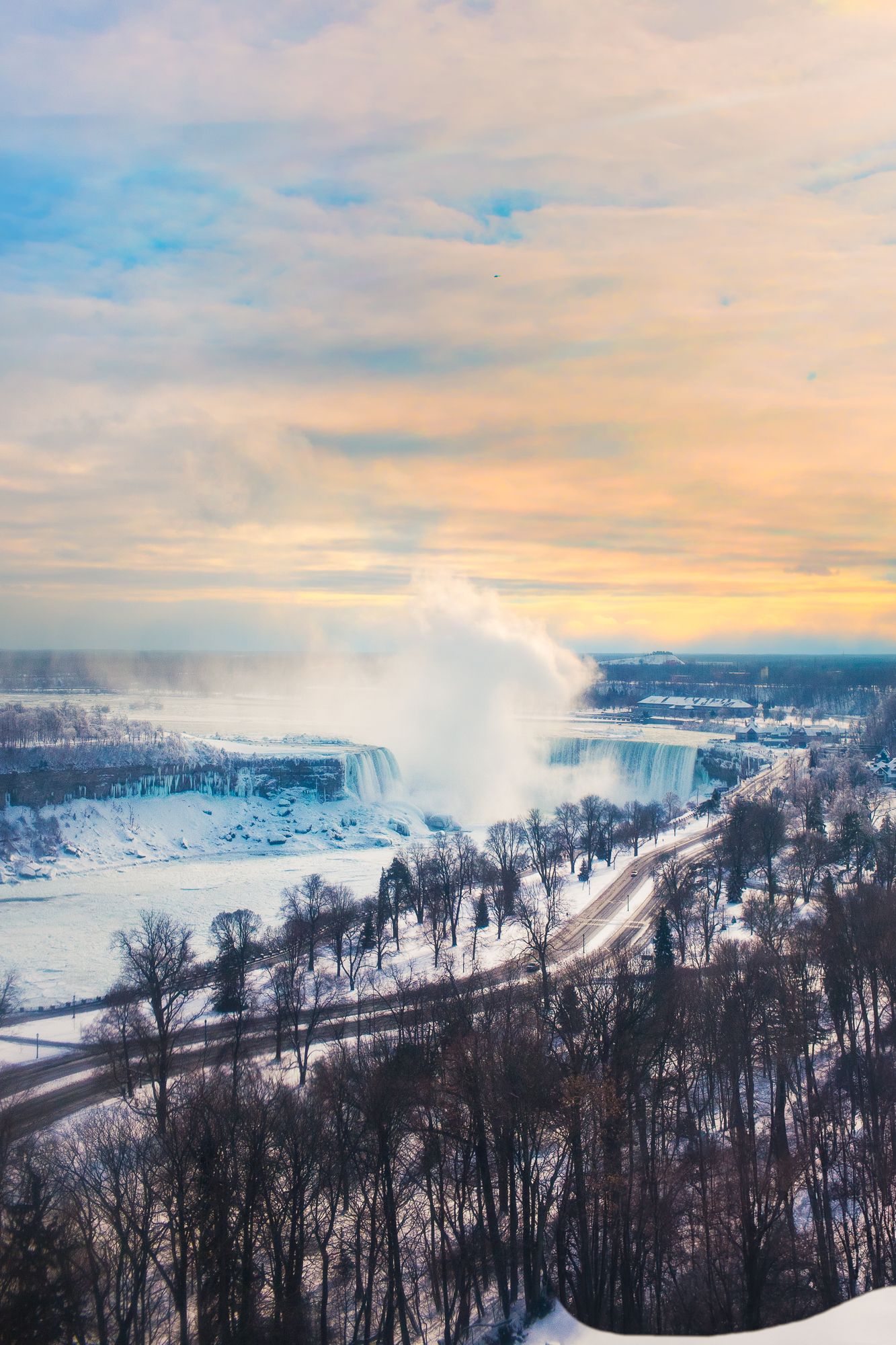Aerial winter view of Niagara Falls with mist rising against a sunset sky and snow-covered trees below.