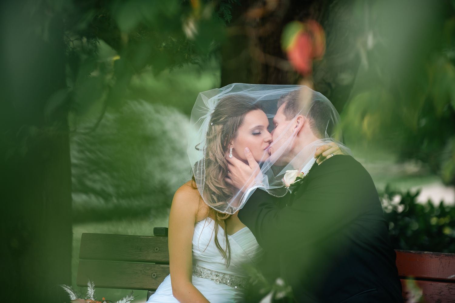 Romantic wedding couple shares an intimate kiss on a garden bench with soft green bokeh background.