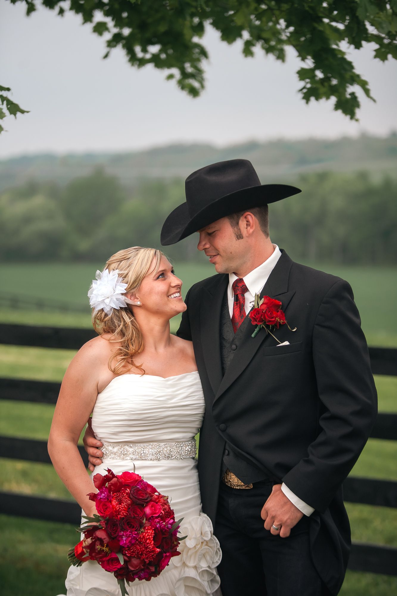 Country western themed wedding couple pose together by wooden fence with red bouquet and cowboy attire.