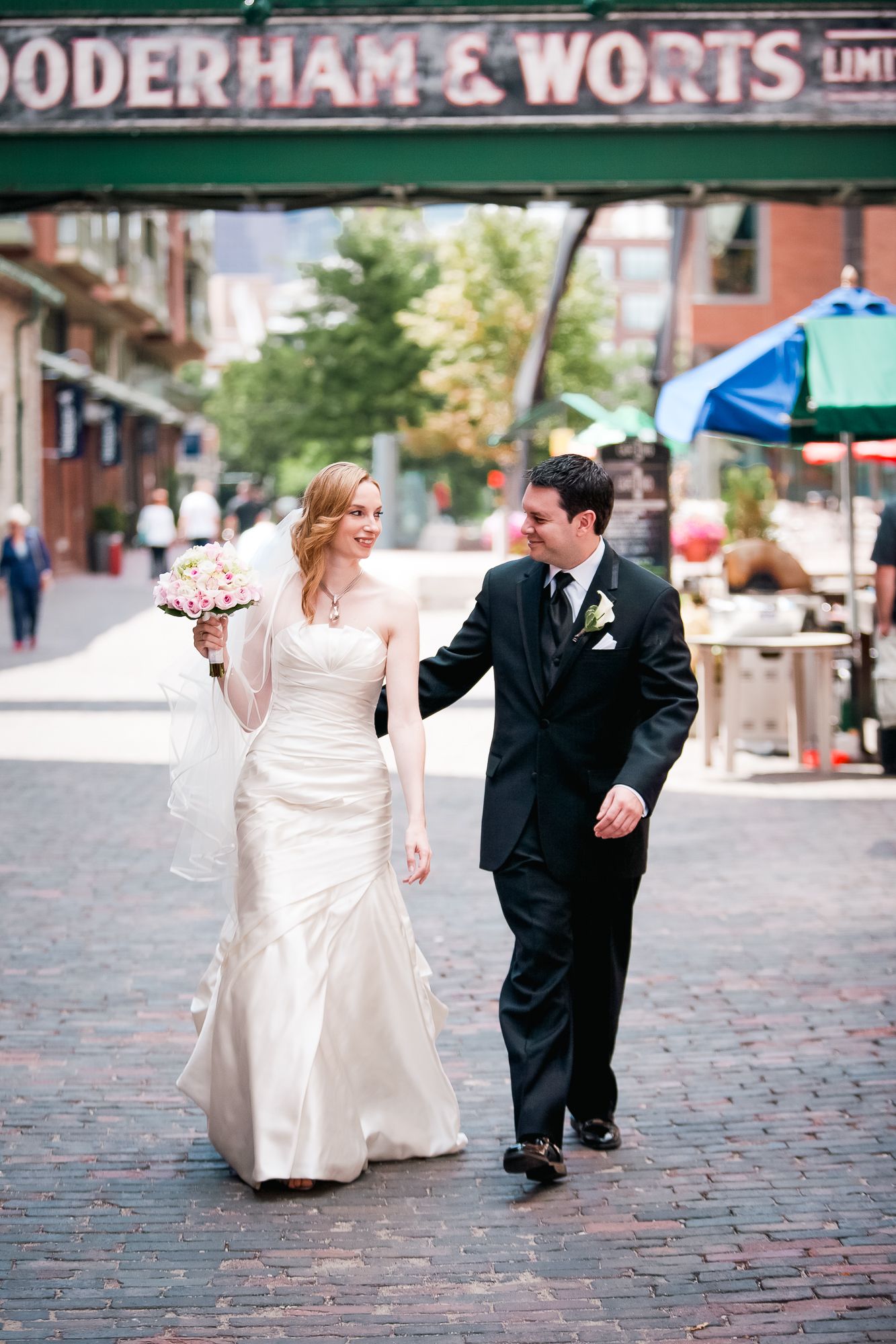 A couple in wedding attire walk along cobblestone streets in front of historic Goderham & Worts building in Toronto.