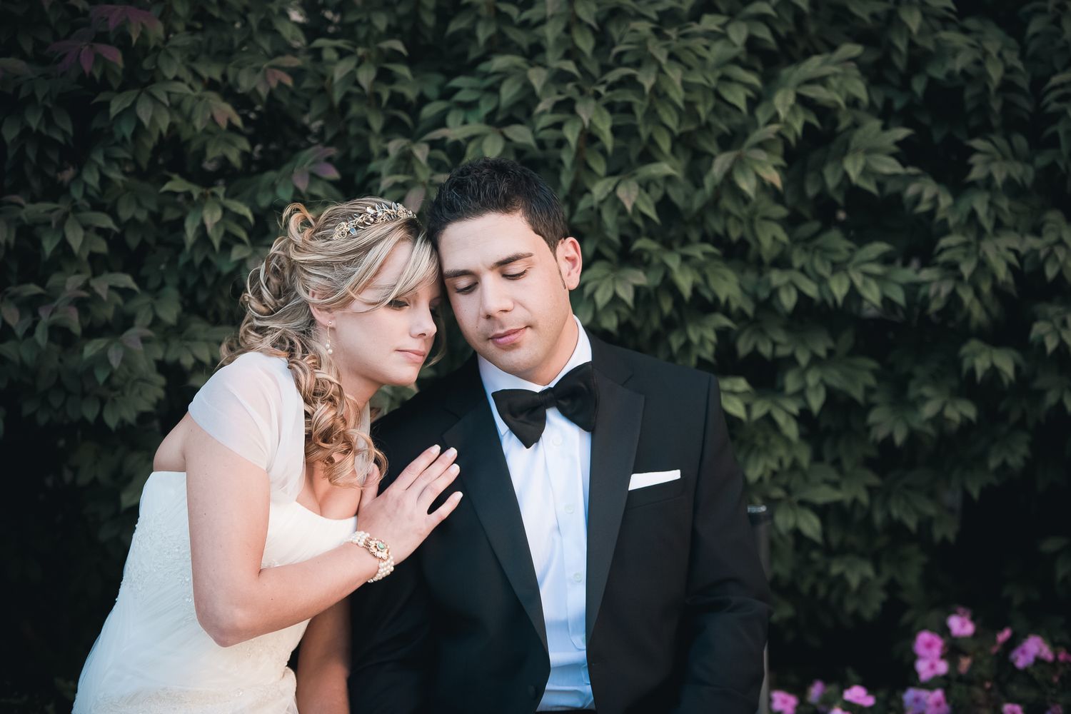 A tender moment between a bride and groom sitting together outdoors with green foliage and pink flowers in the background.