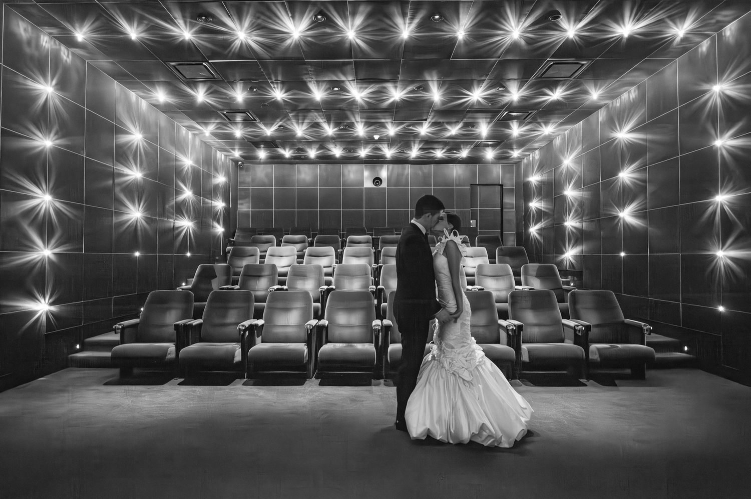 Artistic black and white wedding photo in an empty theater with dramatic starburst lighting effects and rows of seats.