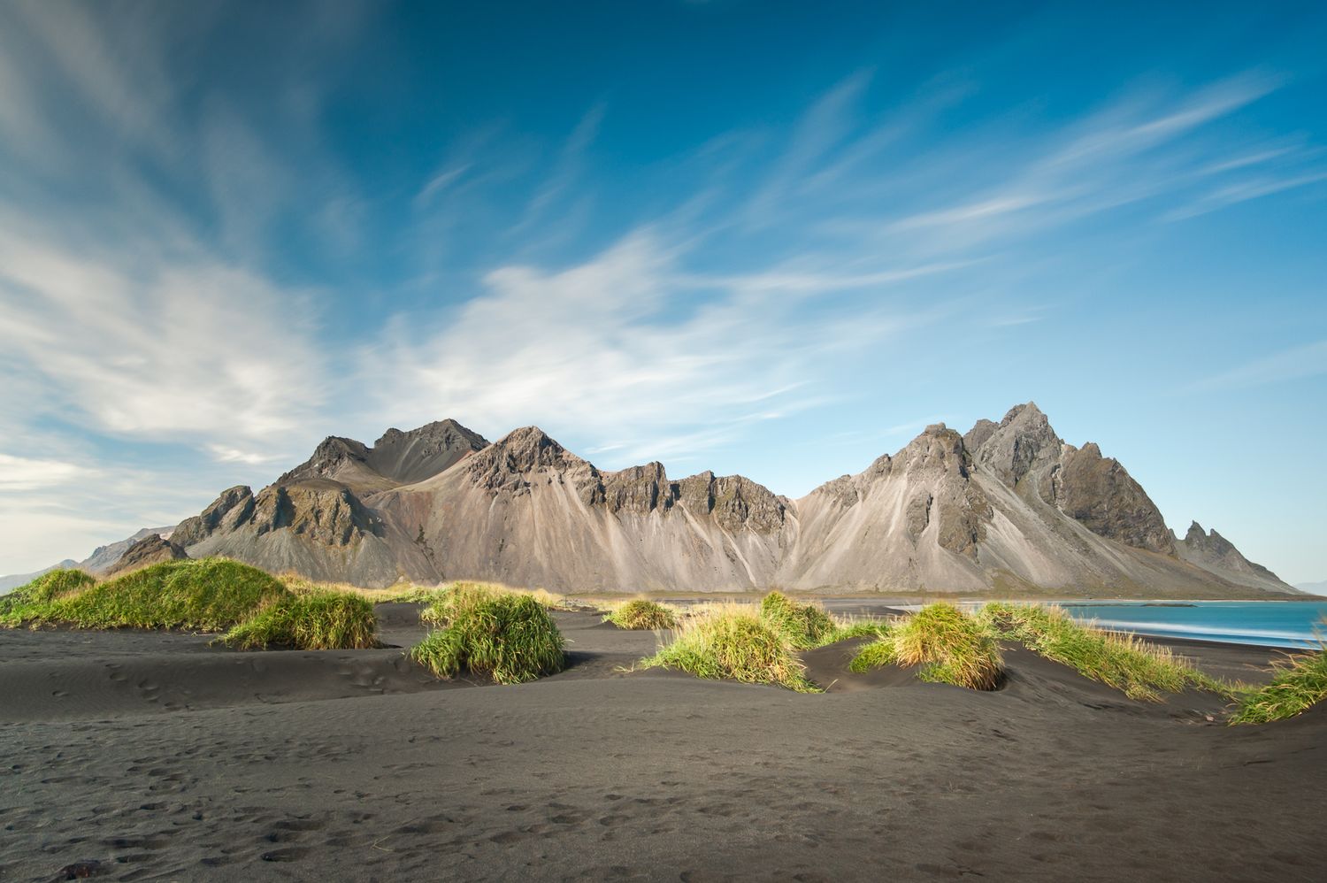 Dramatic view of Vestrahorn mountain range overlooking black sand beach in Iceland under blue sky and wispy clouds.