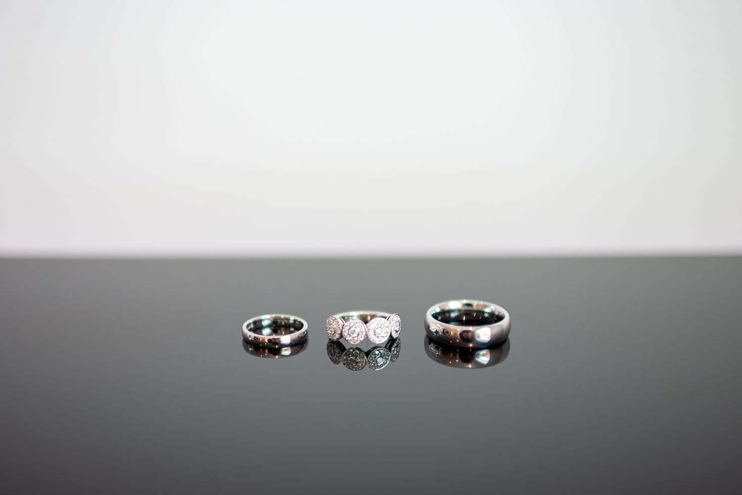 Three elegant rings displayed in a row on a dark reflective surface against a light background.