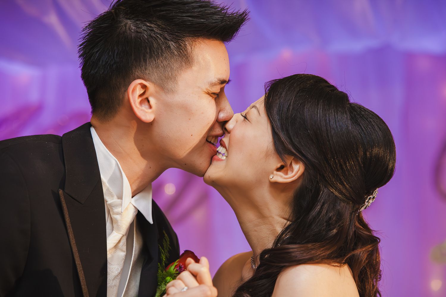 A romantic couple in formal attire share an intimate moment against a purple-lit background with soft bokeh effects.