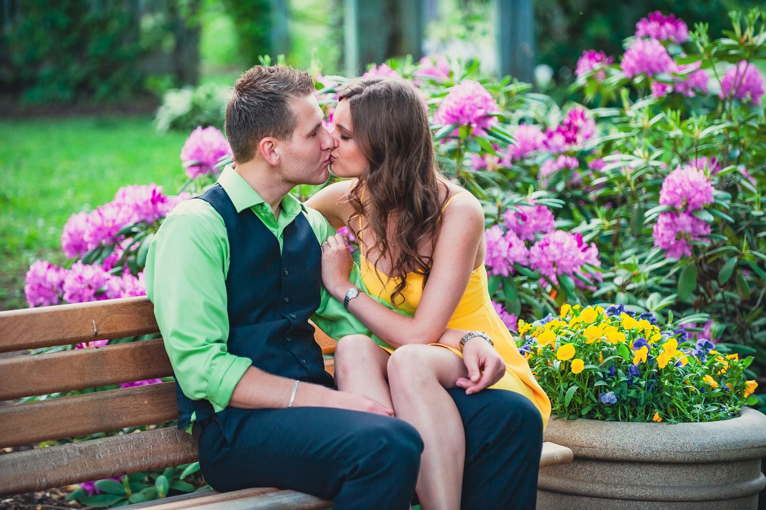 A romantic moment shared on a wooden bench surrounded by vibrant pink flowers and yellow blooms in a garden setting.