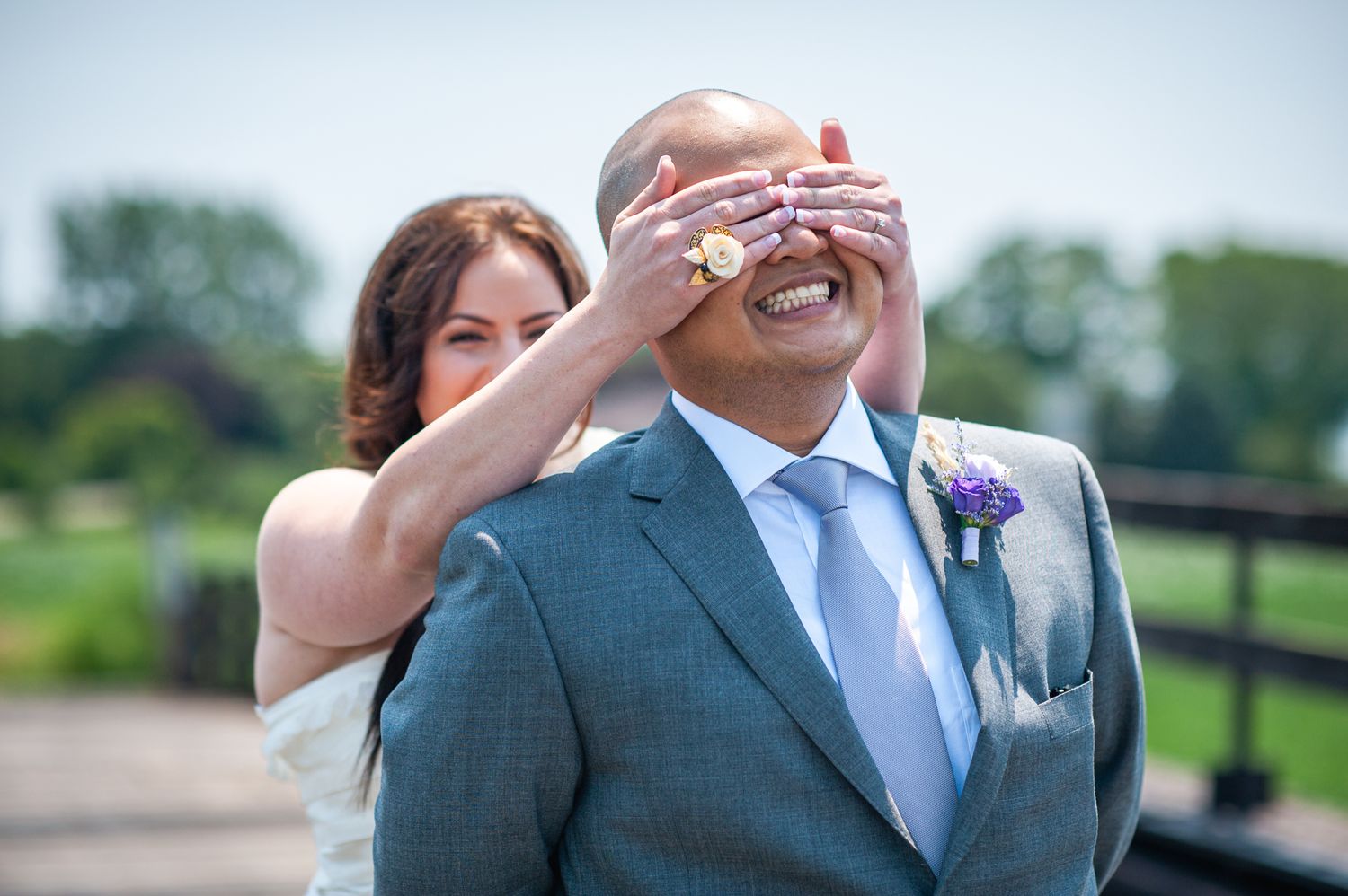 A playful moment between a couple on their wedding day, with the bride covering the groom's eyes outdoors.