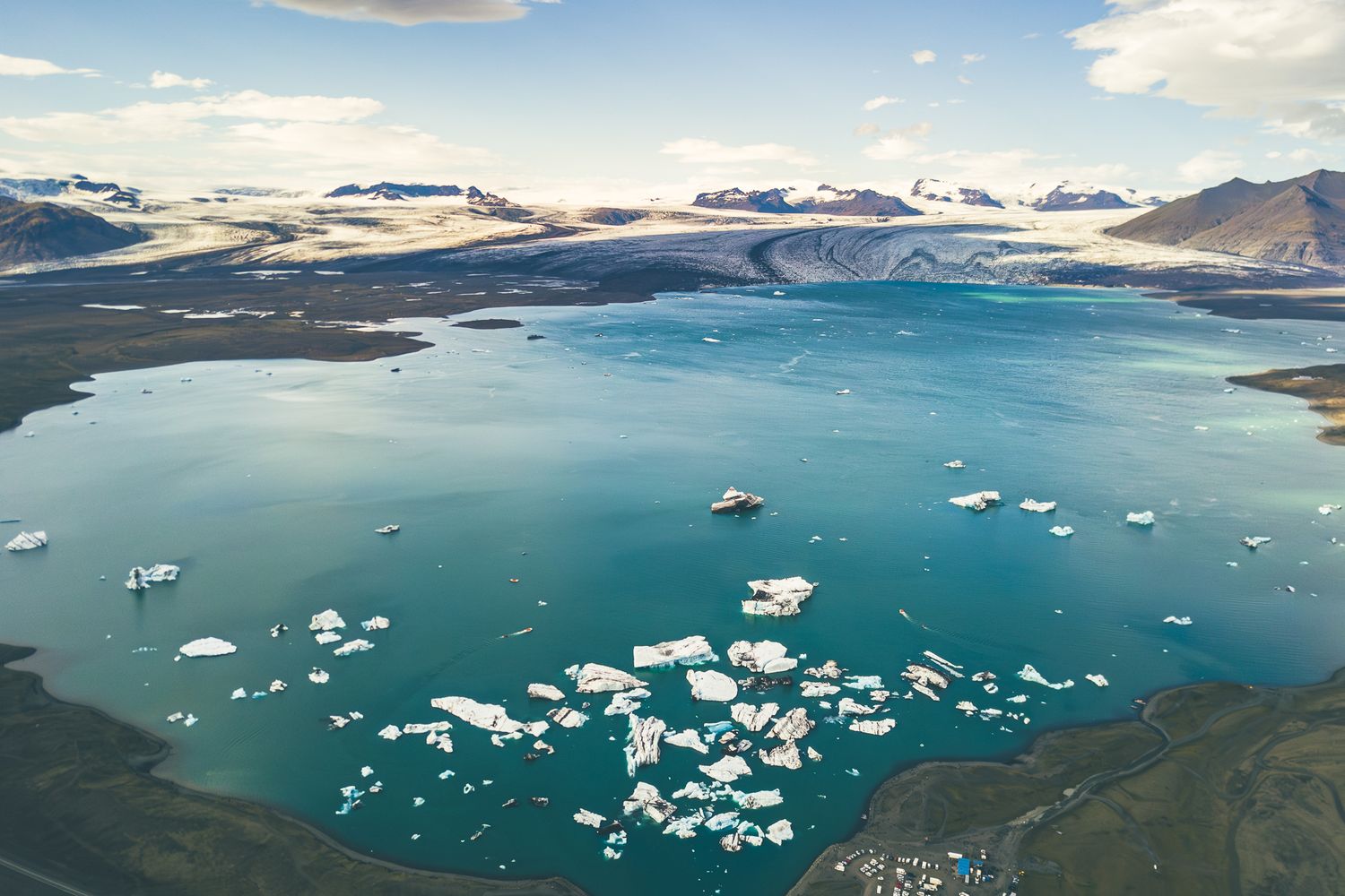 Aerial view of turquoise glacial lake with floating icebergs and surrounding mountains under blue sky.