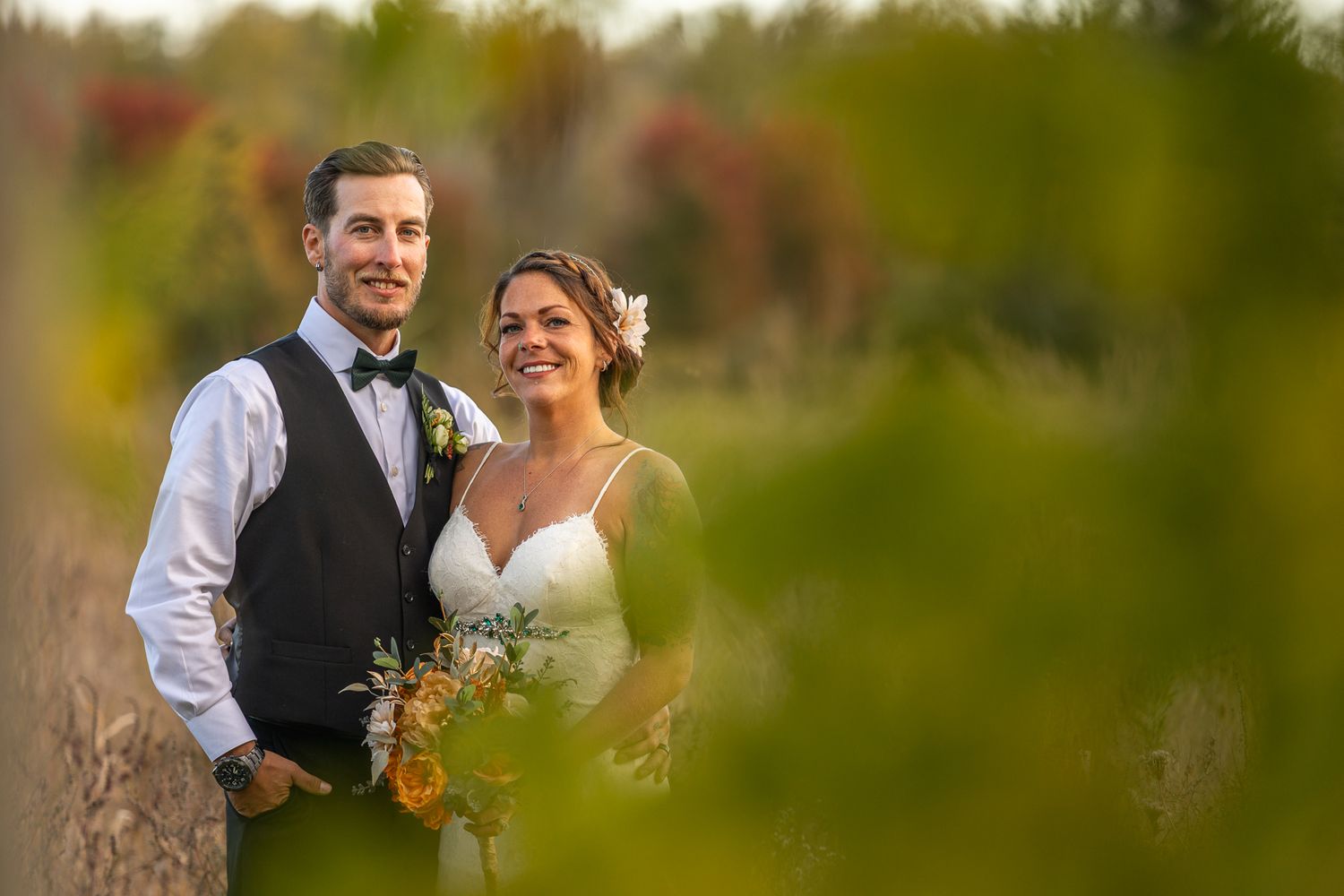 A couple poses together outdoors in wedding attire surrounded by soft bokeh and natural greenery.