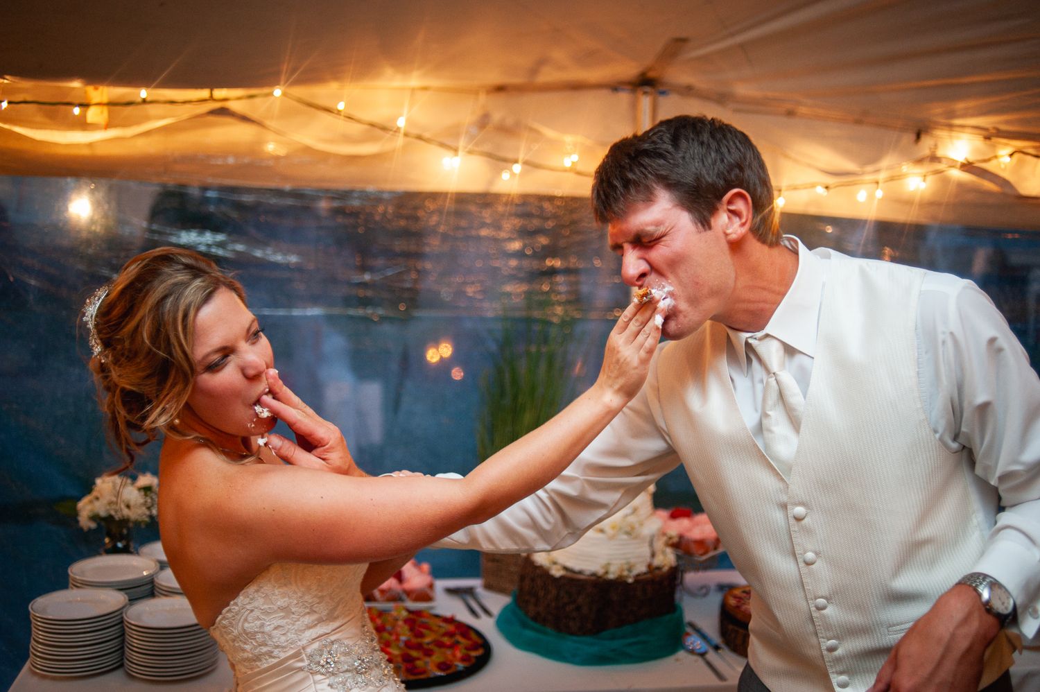 A playful moment between a couple sharing wedding cake at their reception with twinkle lights in the background.