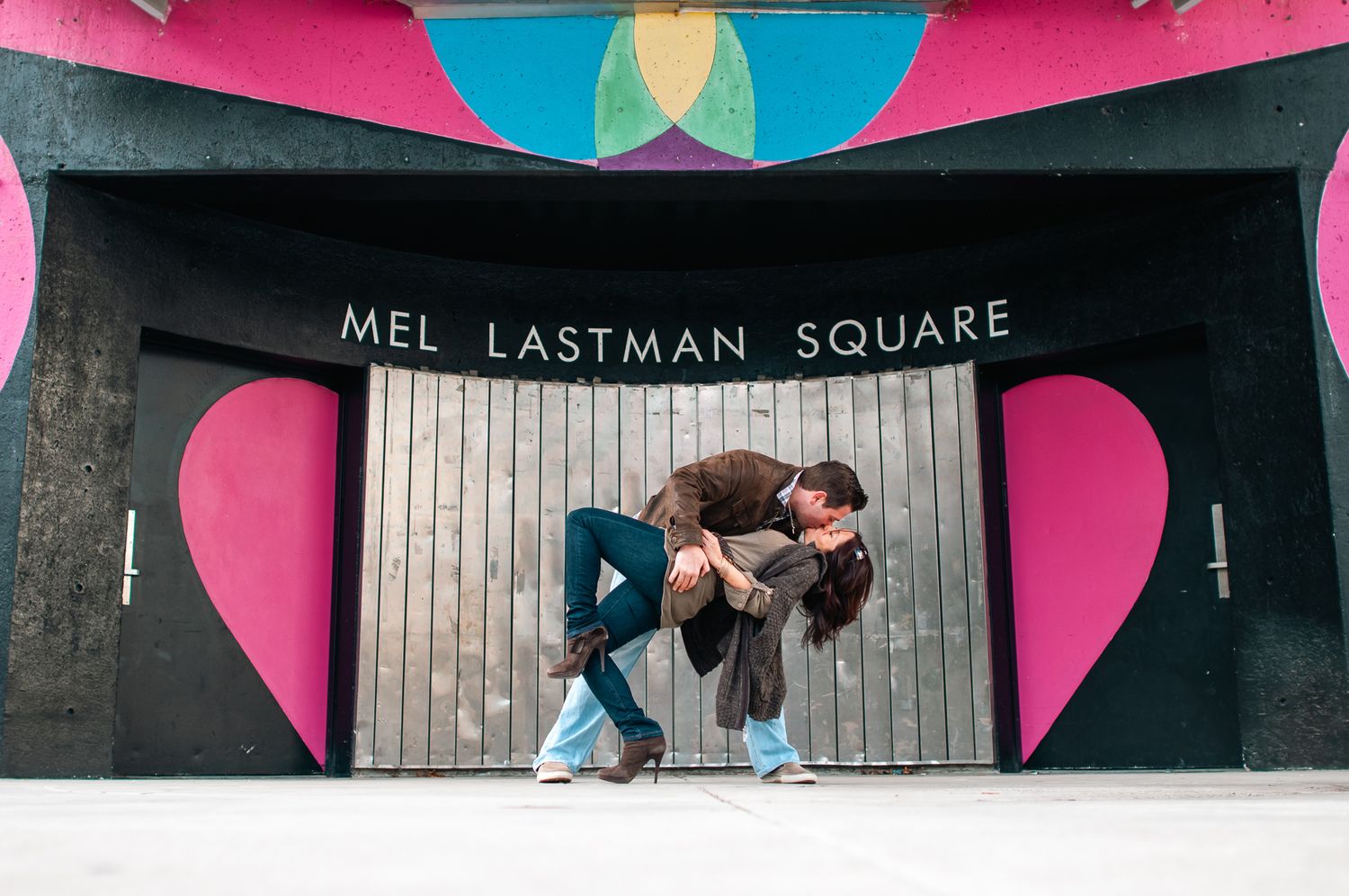 Two dancers perform under a pink and blue urban archway labeled Mel Lastman Square.
