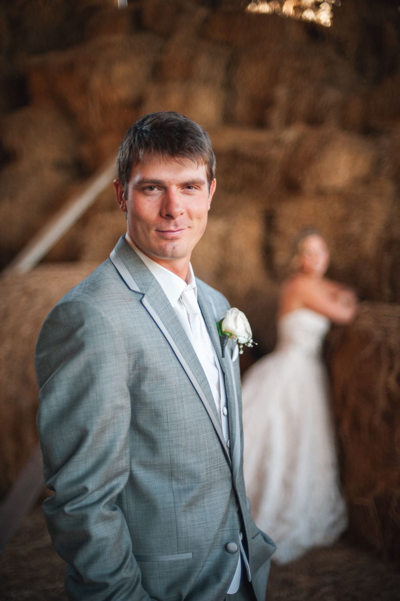 A groom in a light grey suit with boutonniere poses for a wedding photo with a bride visible in the background.