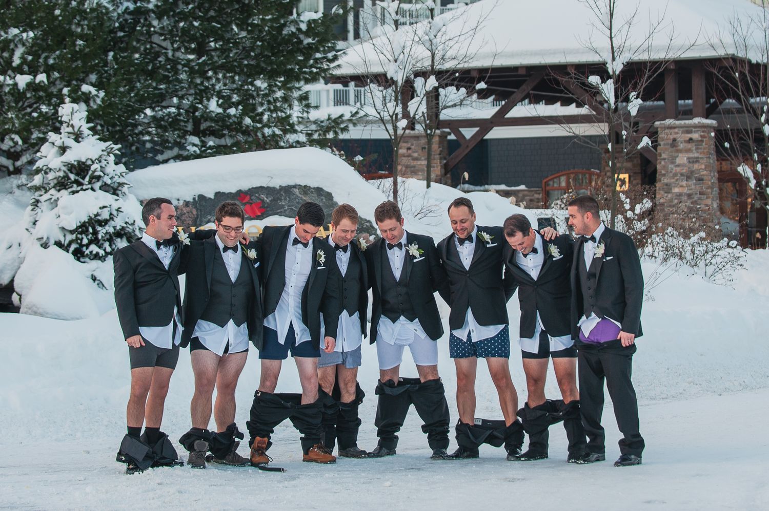 A group in matching formal attire with shorts pose together in the snow for a winter wedding photo.
